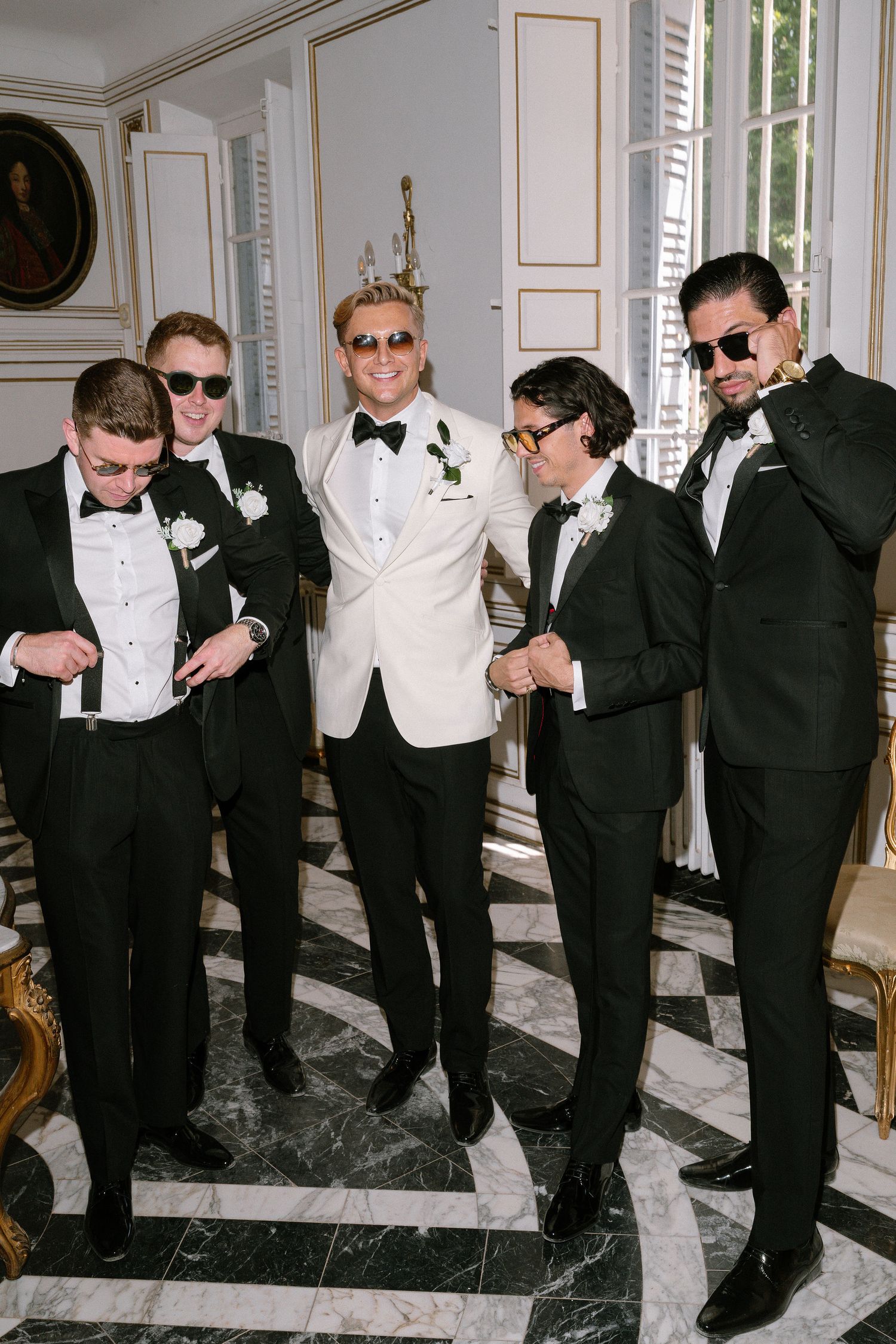 Groom in white tuxedo jacket poses with four groomsmen in black suits and sunglasses in ornate marble interior.