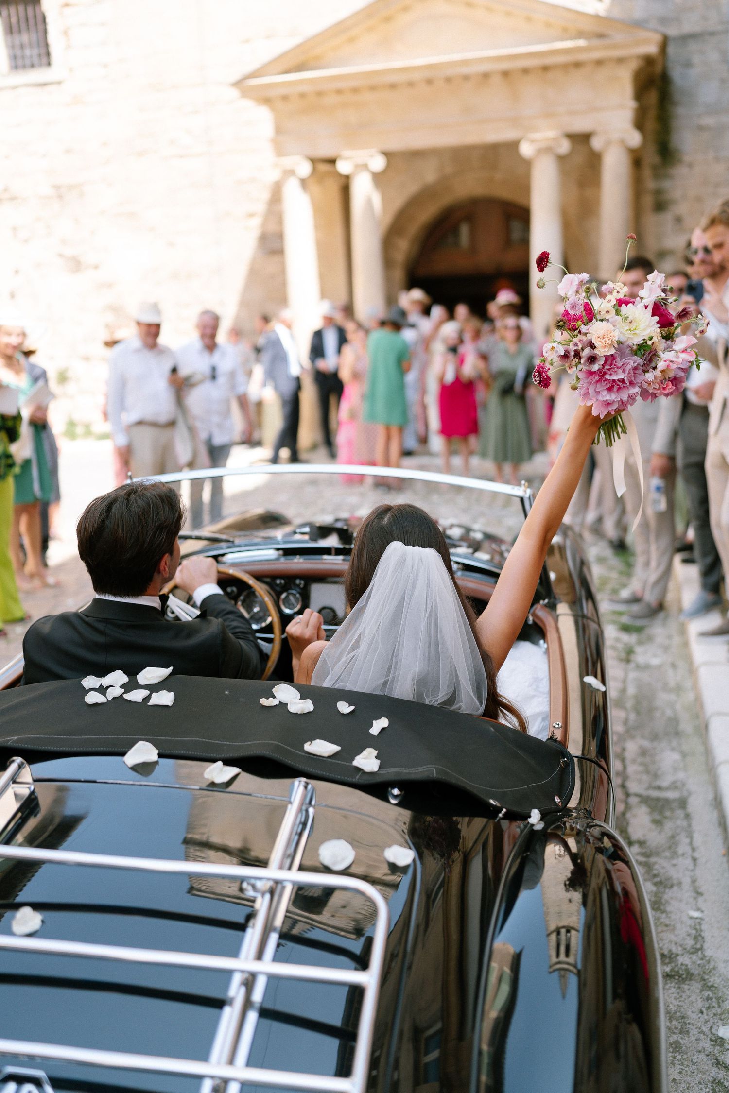 Wedding party waves from decorated vintage cars during a celebratory departure.