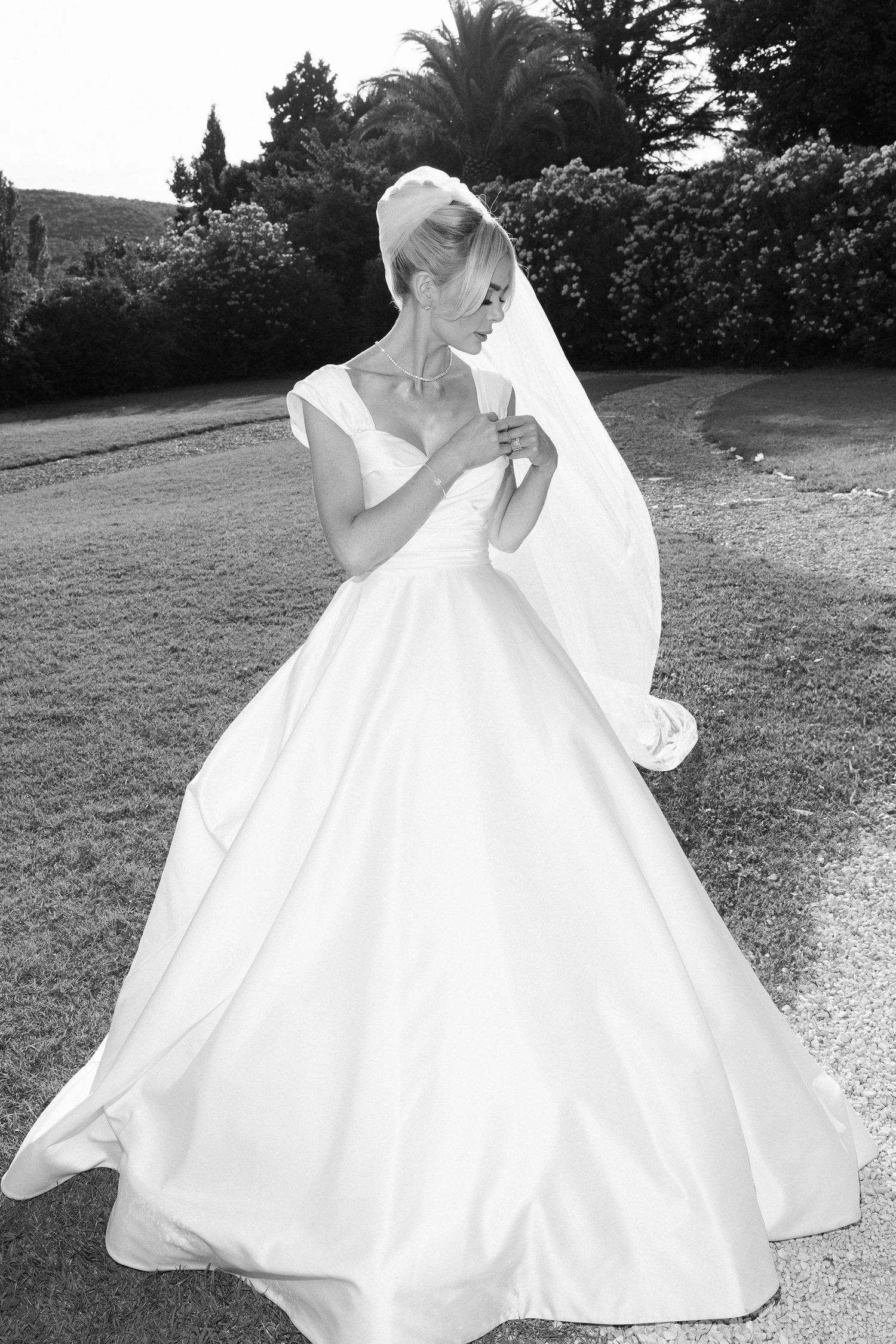 Elegant bride in a flowing white ball gown and veil poses outdoors in a lush garden setting.