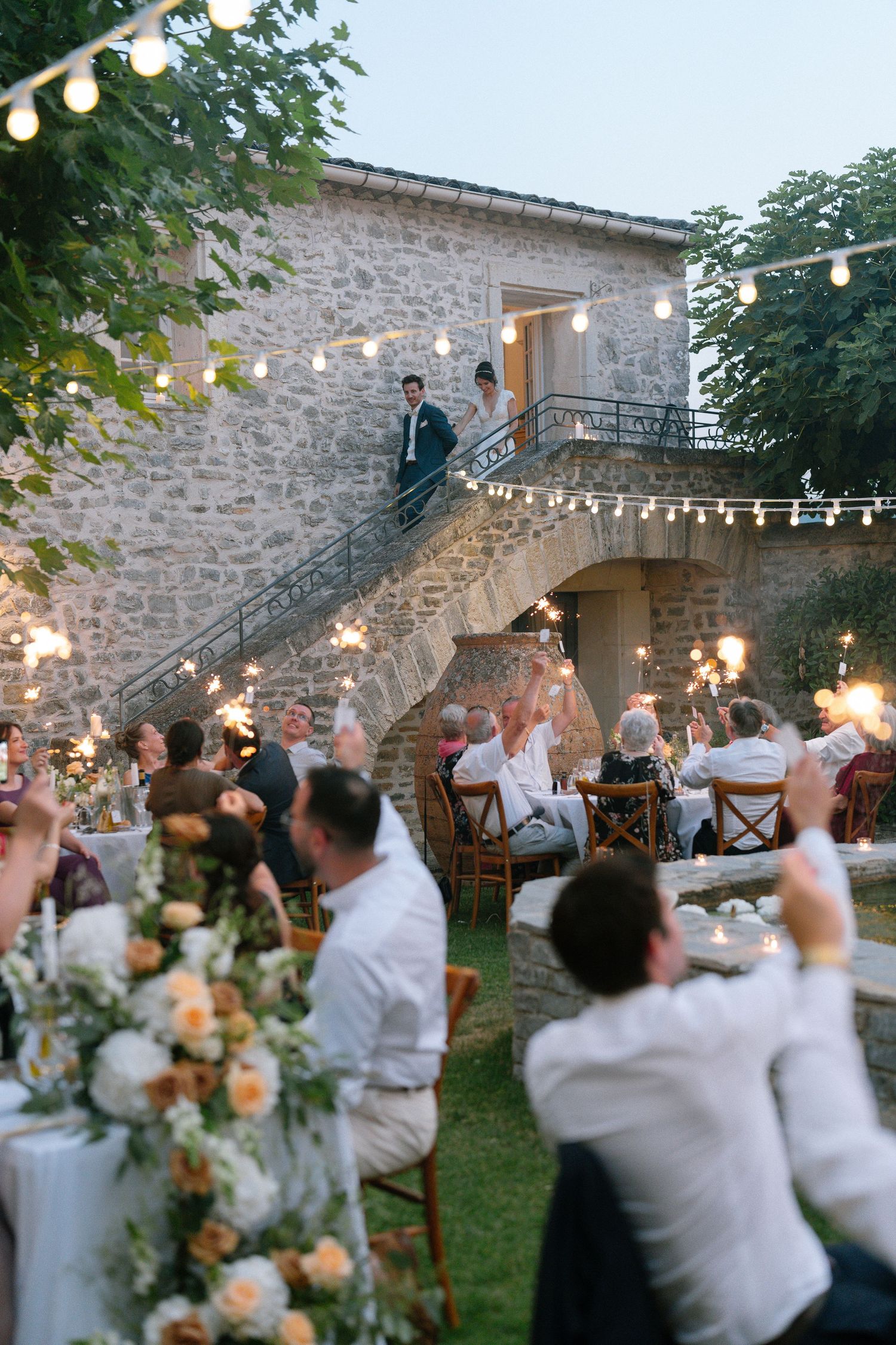Wedding guests raise their glasses in celebration at an outdoor reception with string lights and stone walls.