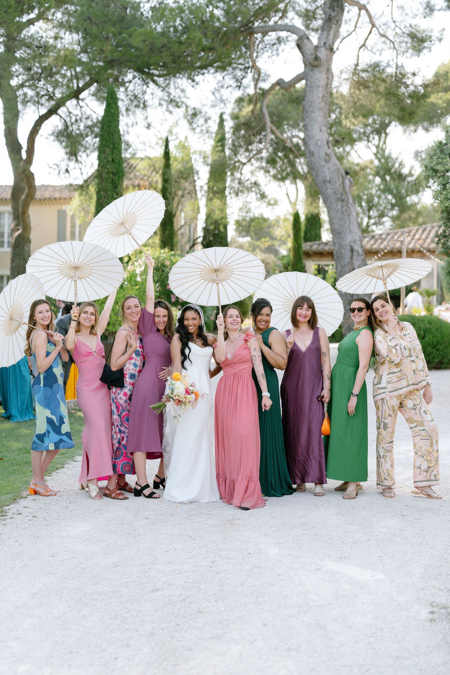 Wedding party holding white parasols poses outdoors in colorful dresses on a sunny day.