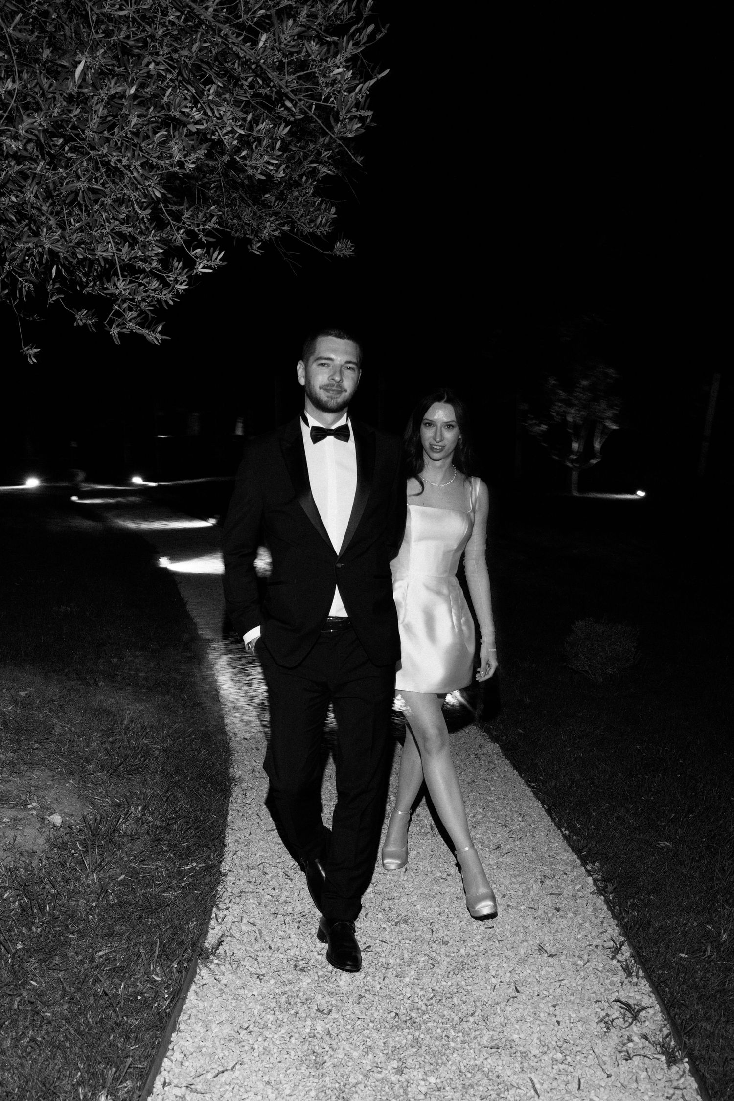 Elegant couple in black and white photo, man in tuxedo and woman in white mini dress walking at night.