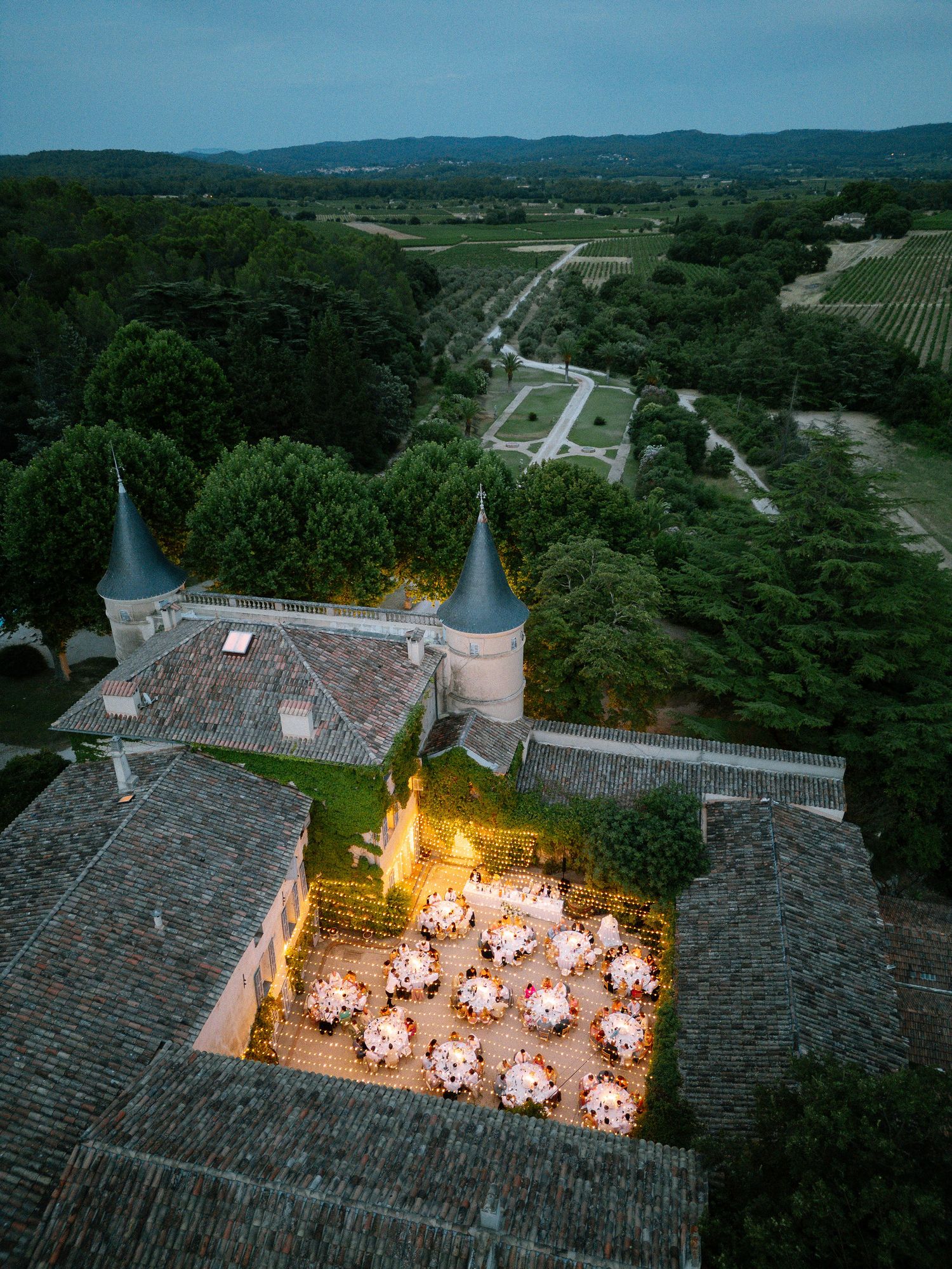 Aerial view of a French chateau courtyard with illuminated reception tables at dusk.