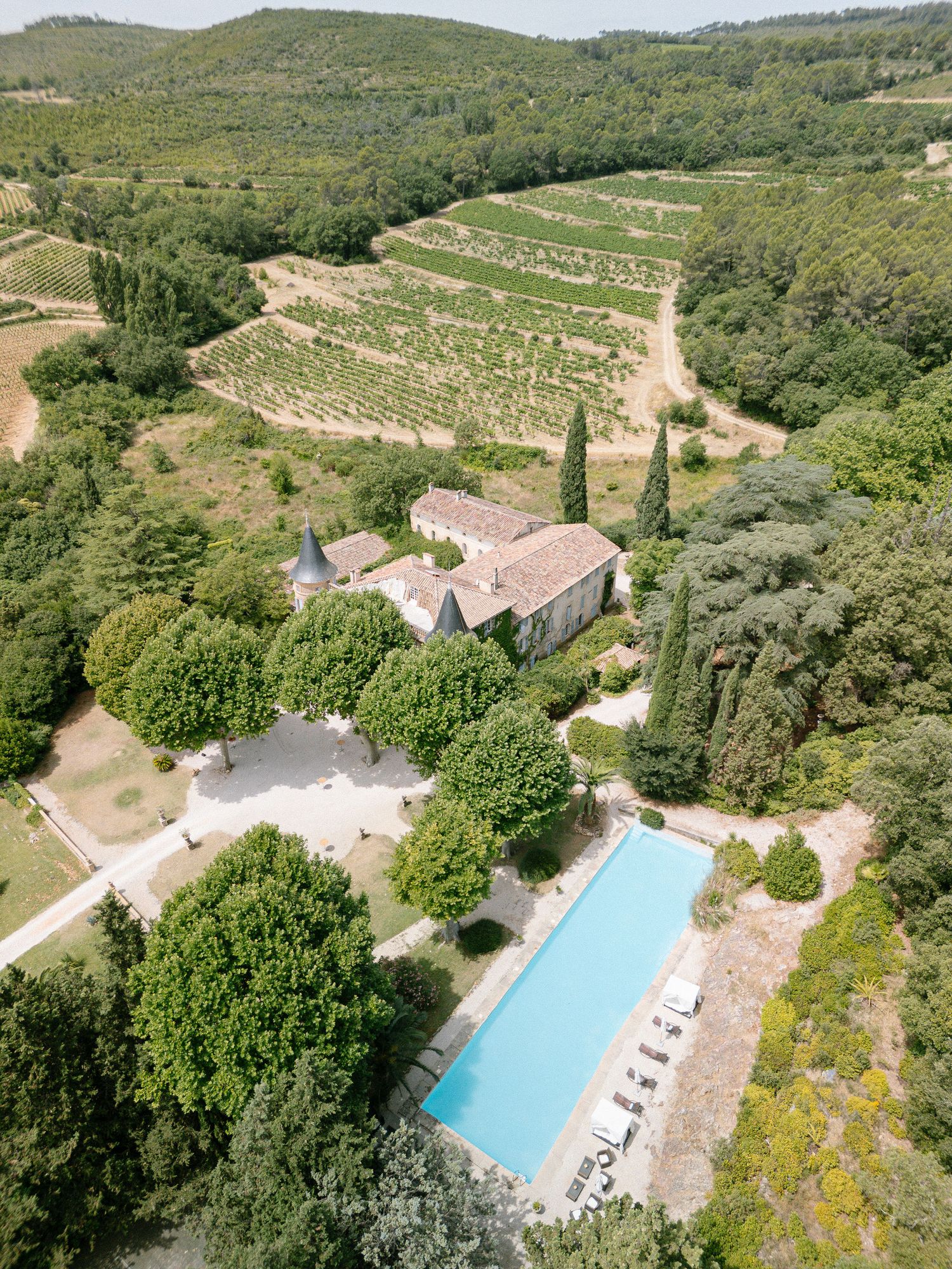 Aerial view of a French countryside estate with a rectangular pool, stone buildings, cypress trees, and vineyard hillsides.