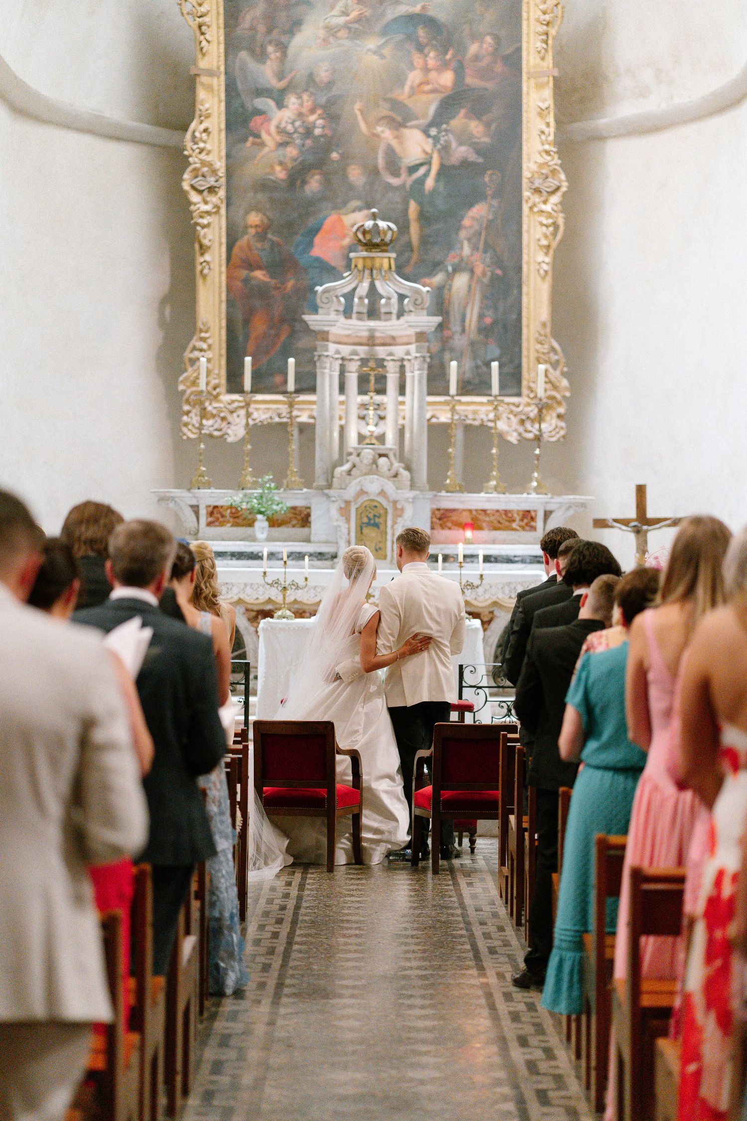 Bride and groom at church altar during wedding ceremony with guests standing in ornate chapel with baroque painting.