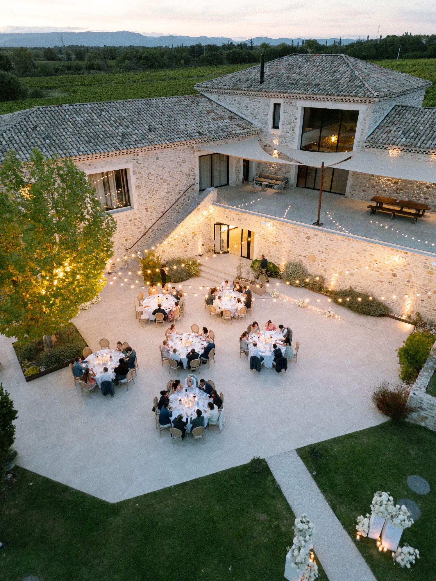 Aerial view of outdoor wedding reception with stone courtyard and illuminated dining tables at dusk.