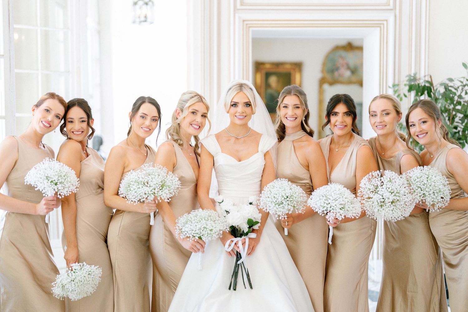 A bride with eight bridesmaids in champagne gowns holding white floral bouquets at a wedding.