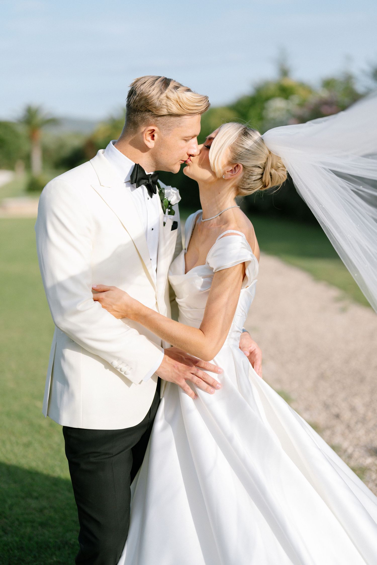 Elegant wedding couple embracing outdoors with flowing veil in natural light.