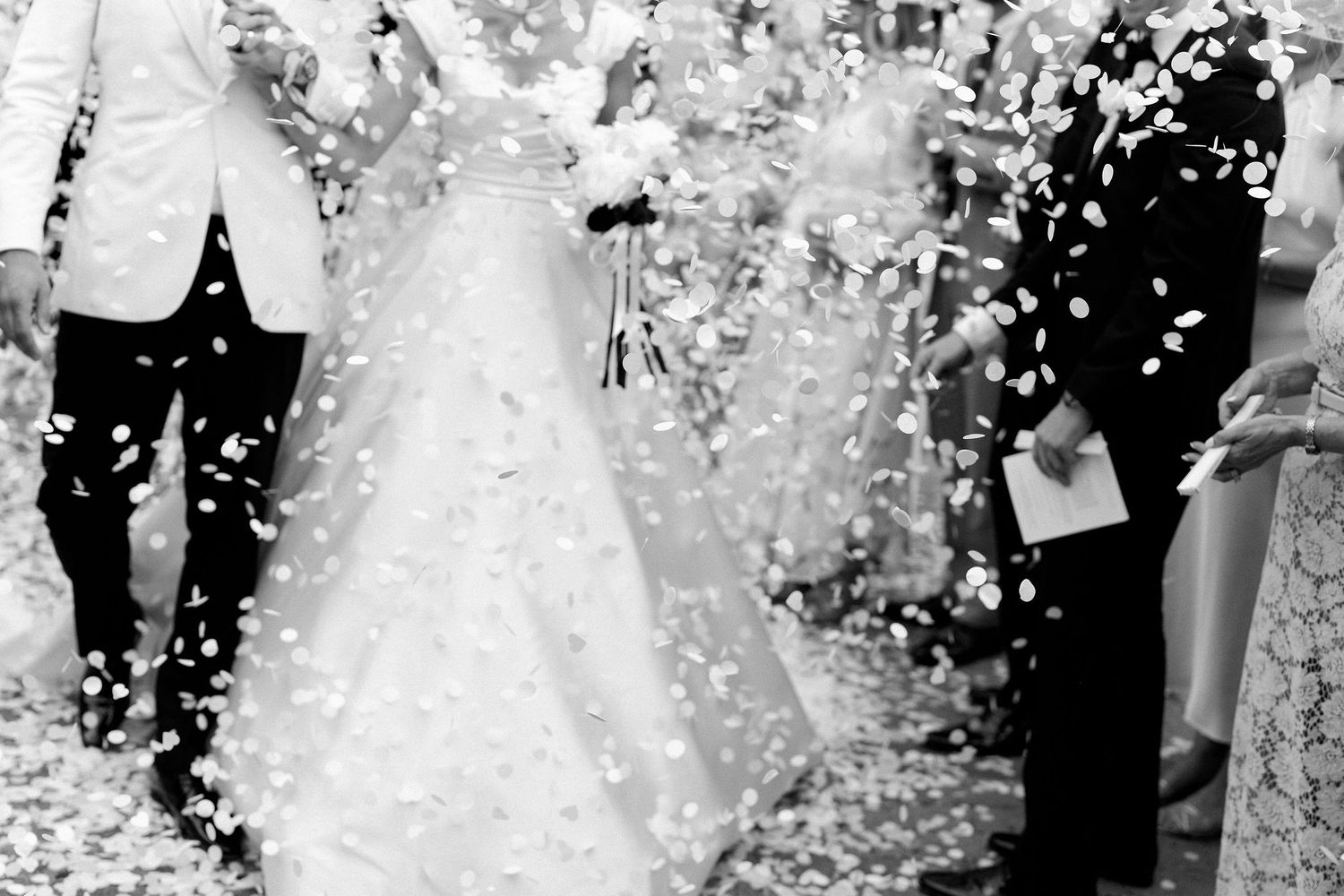 Black and white photo of bride and groom walking through confetti shower at outdoor wedding ceremony.