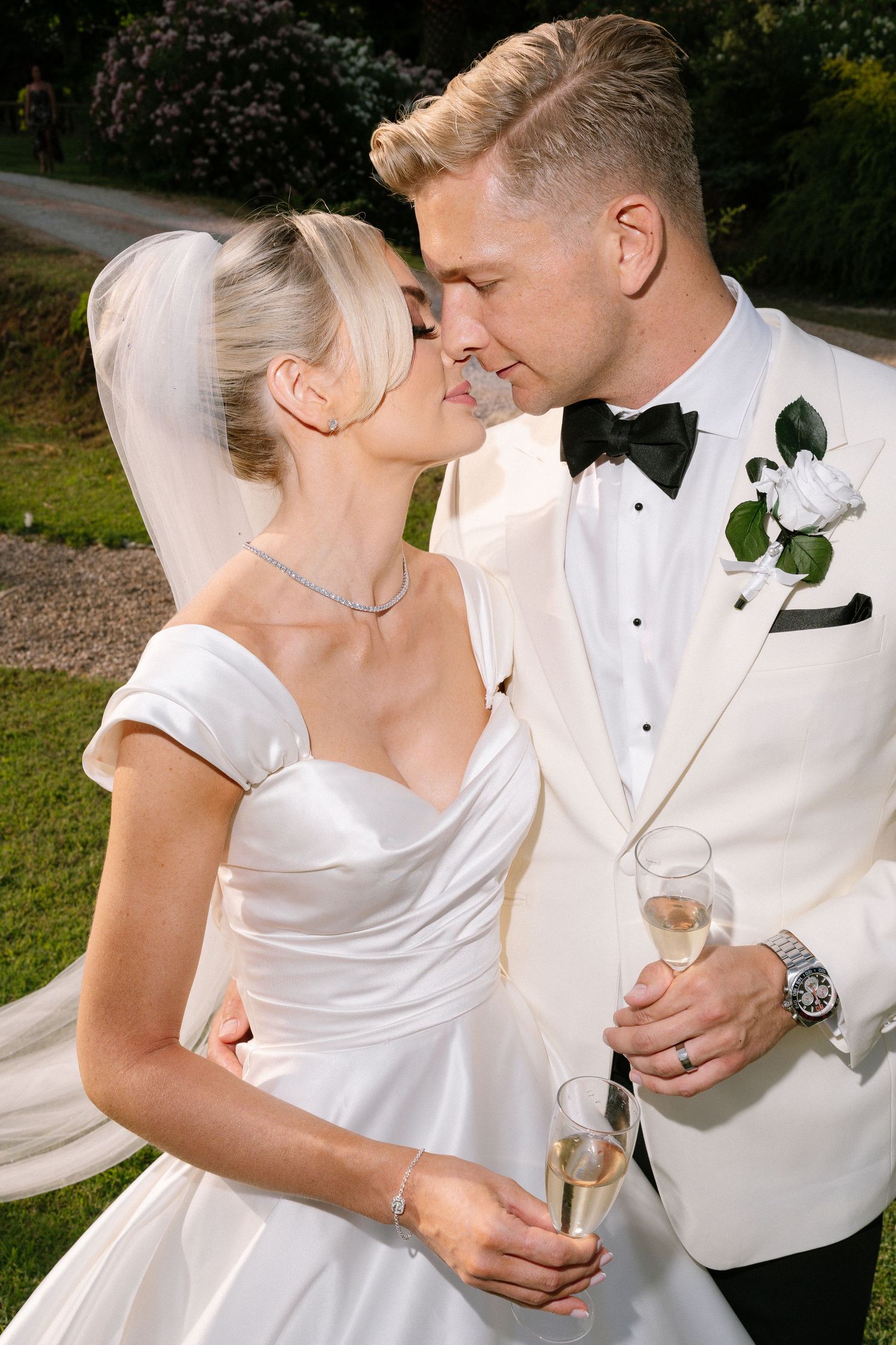 Romantic wedding couple in white attire sharing intimate moments and champagne during outdoor photoshoot.