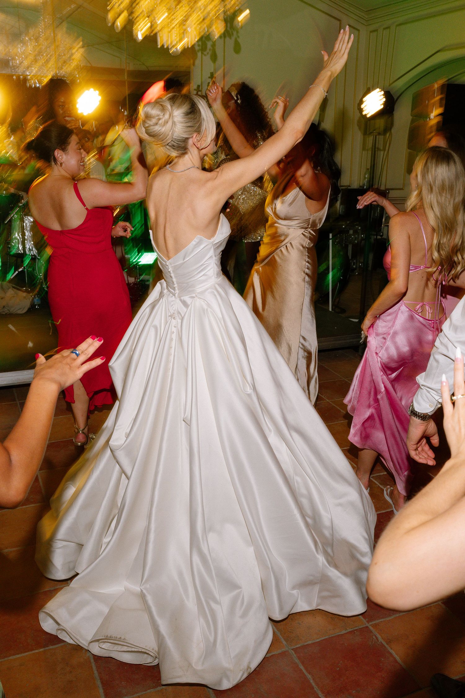 Bride in white ballgown dancing with guests at wedding reception, bridesmaids in pink and red dresses celebrating.