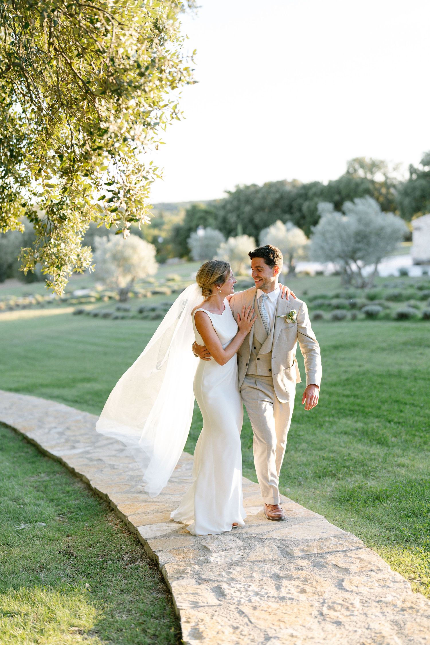 A couple in wedding attire walks along a winding stone path through green grounds.