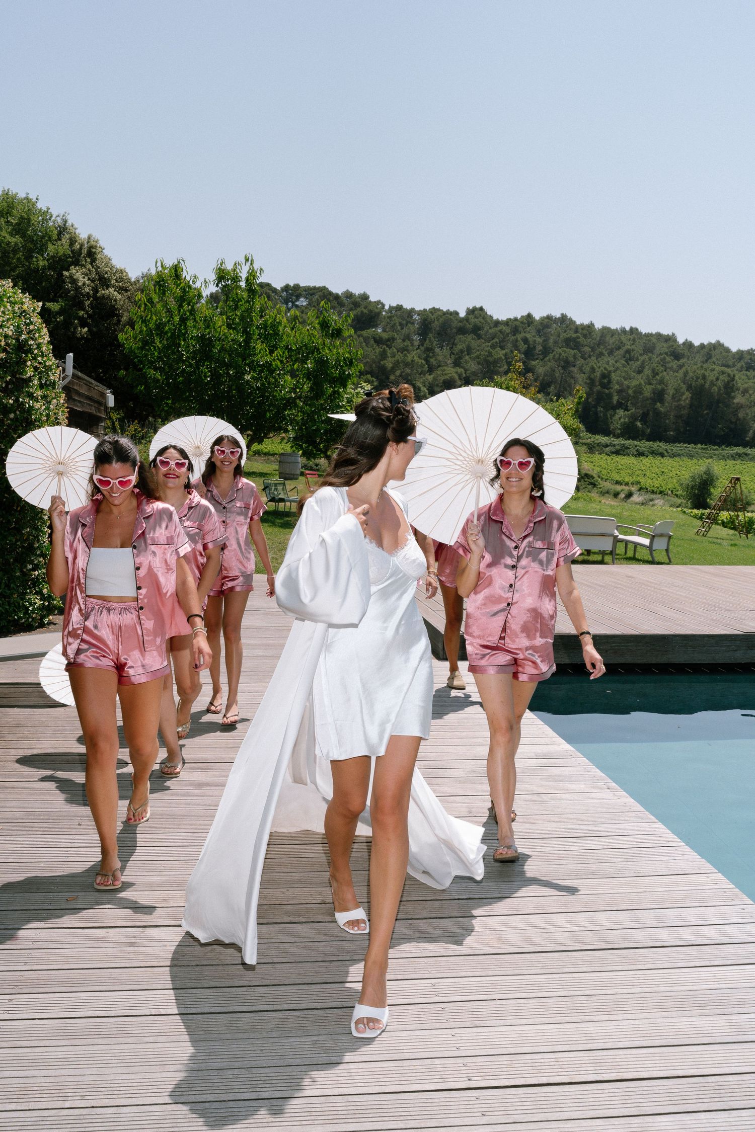 Group walking along poolside deck carrying parasols in coordinated pink outfits.