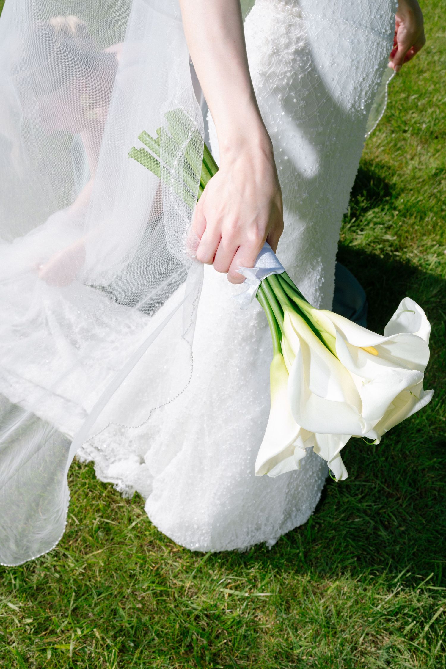 Bride in white lace gown holding elegant calla lily bouquet on green grass on her wedding day.