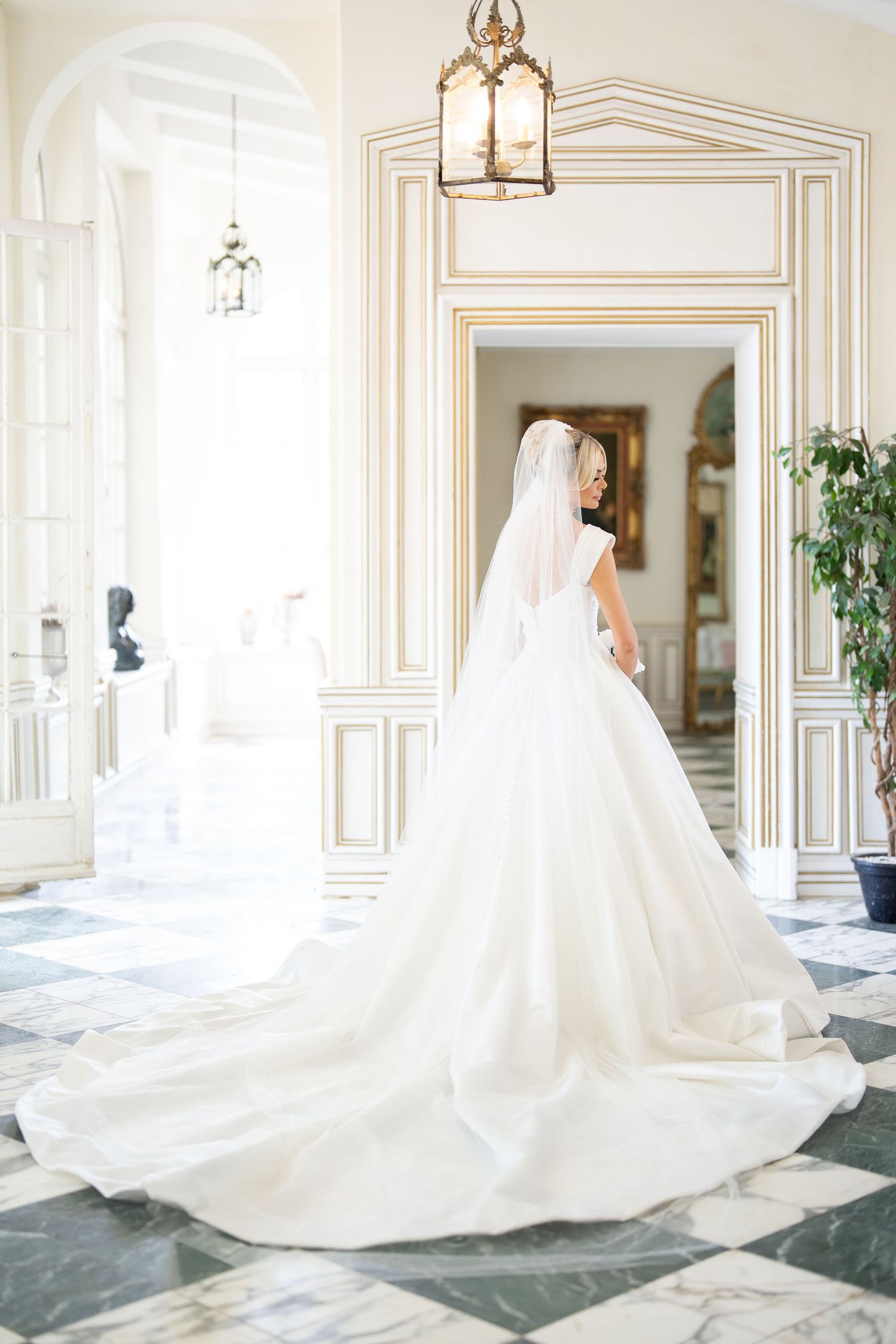 Bride in elegant white ball gown with long train and veil stands in luxurious gold-trimmed venue hallway.