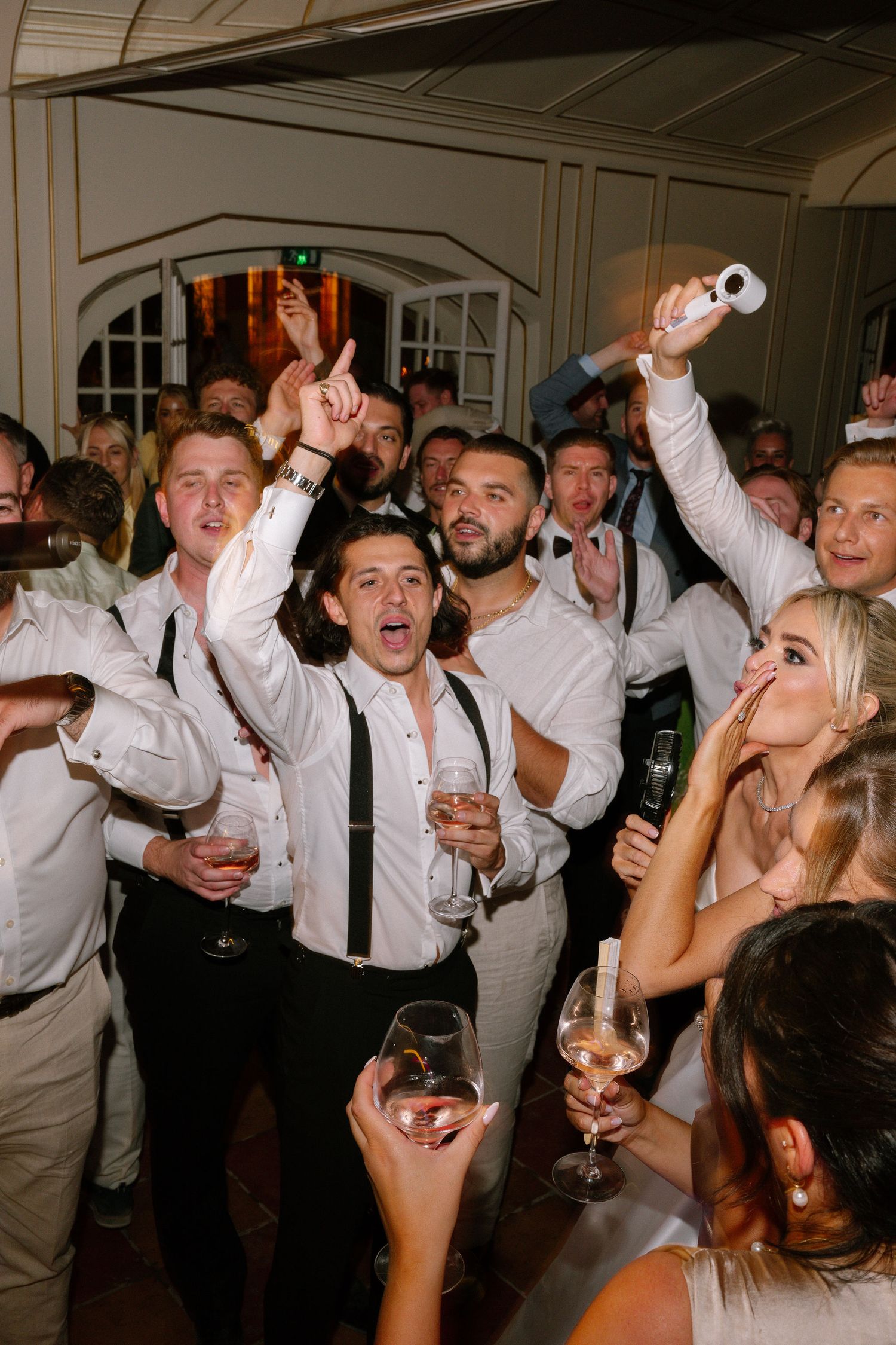 Guests celebrating at a wedding reception, dancing and raising glasses of wine in a lively indoor party atmosphere.
