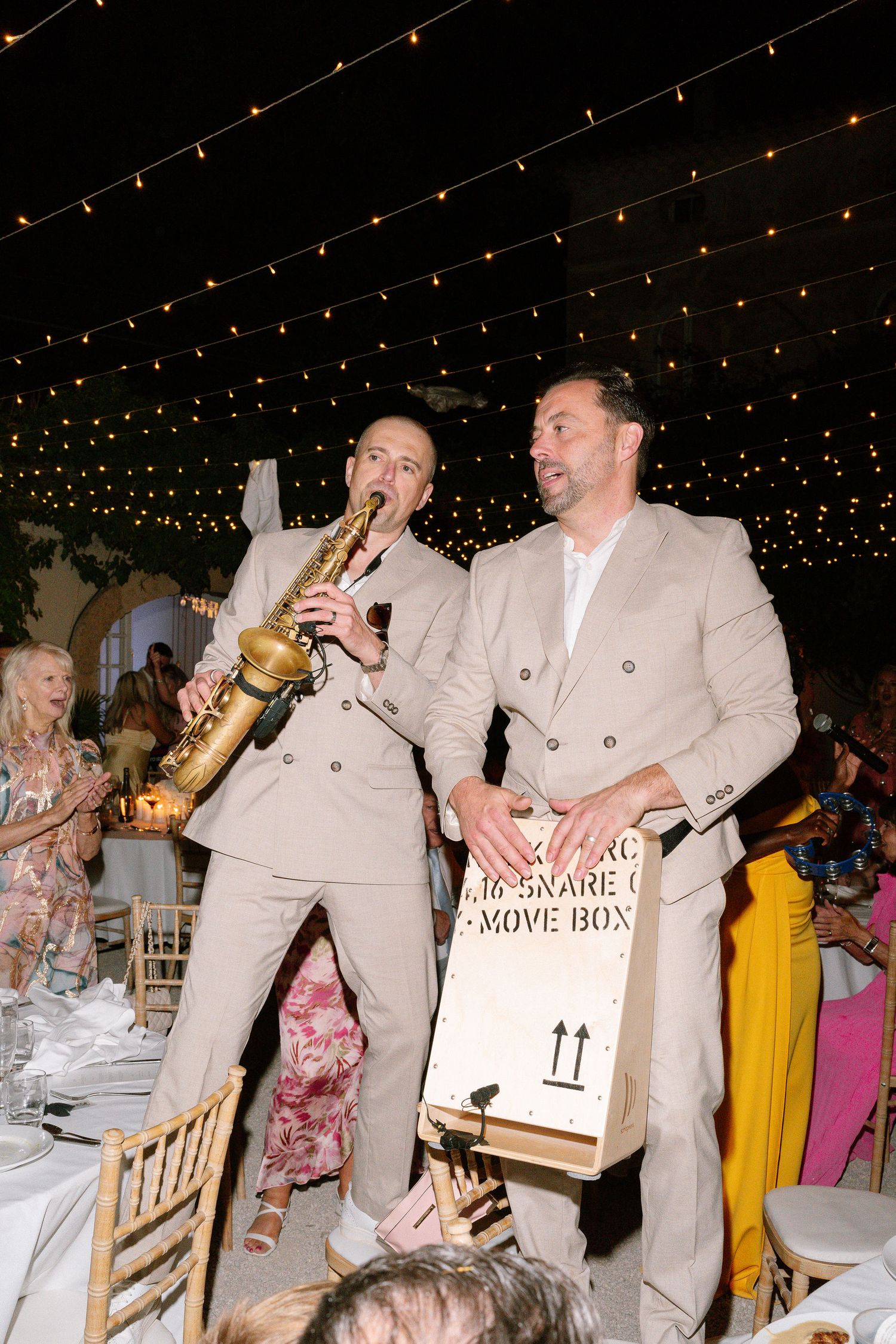 Two musicians in beige suits perform at an outdoor evening event, one playing saxophone and one holding a wooden box.