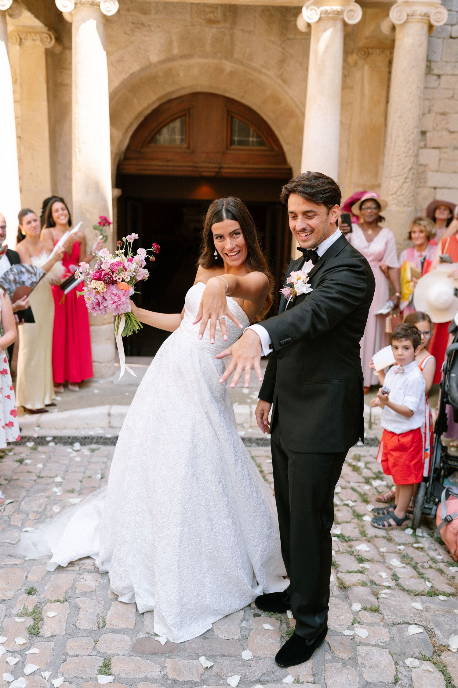 Newlyweds celebrate outside a historic stone building with wedding guests throwing confetti.