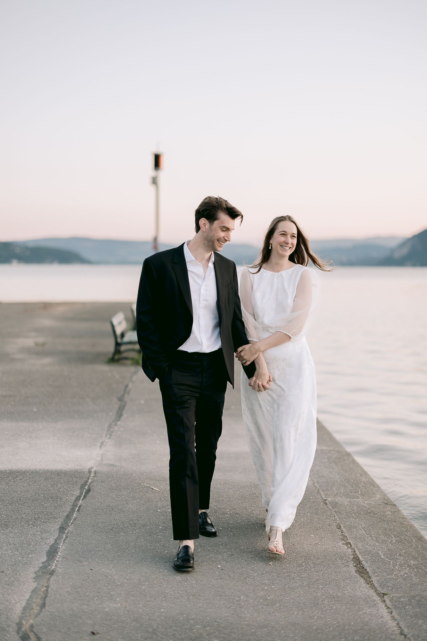 Romantic couple walking along waterfront at sunset in formal attire.