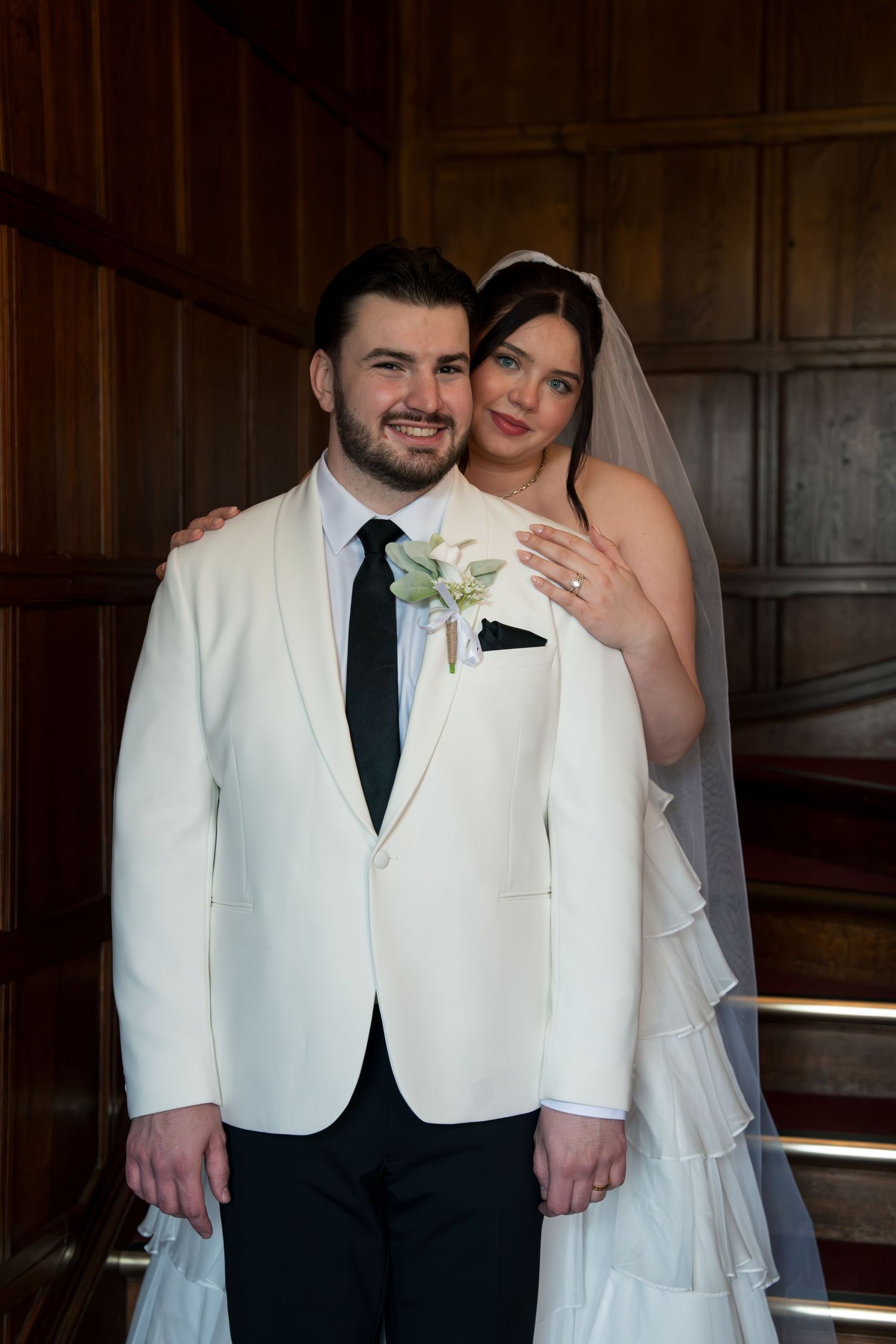 Smiling groom in white tuxedo with boutonniere poses with bride in white gown and veil in elegant wood-paneled room.