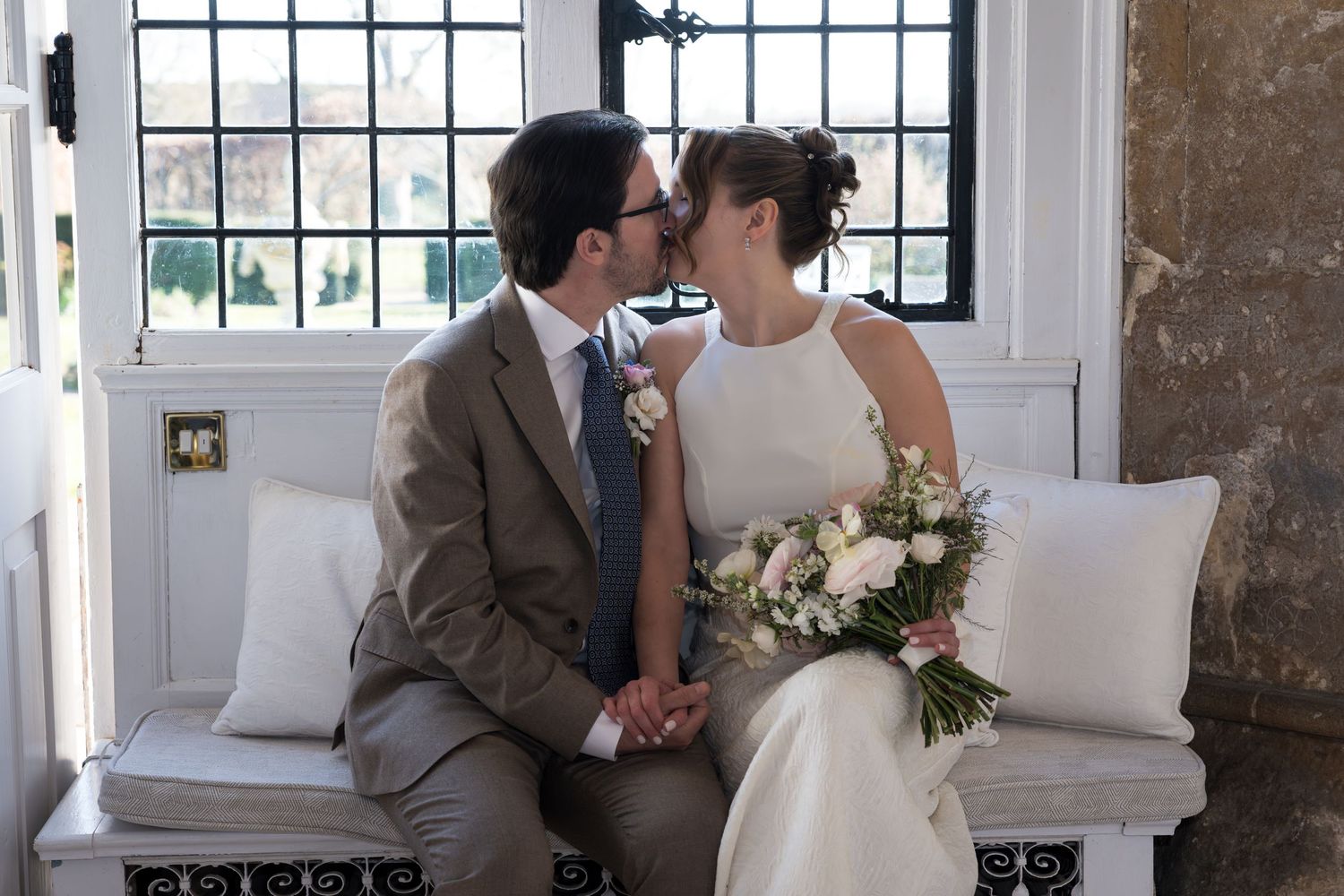 Bride and groom share a kiss on a white bench, bride holding a pink floral bouquet near elegant windows.