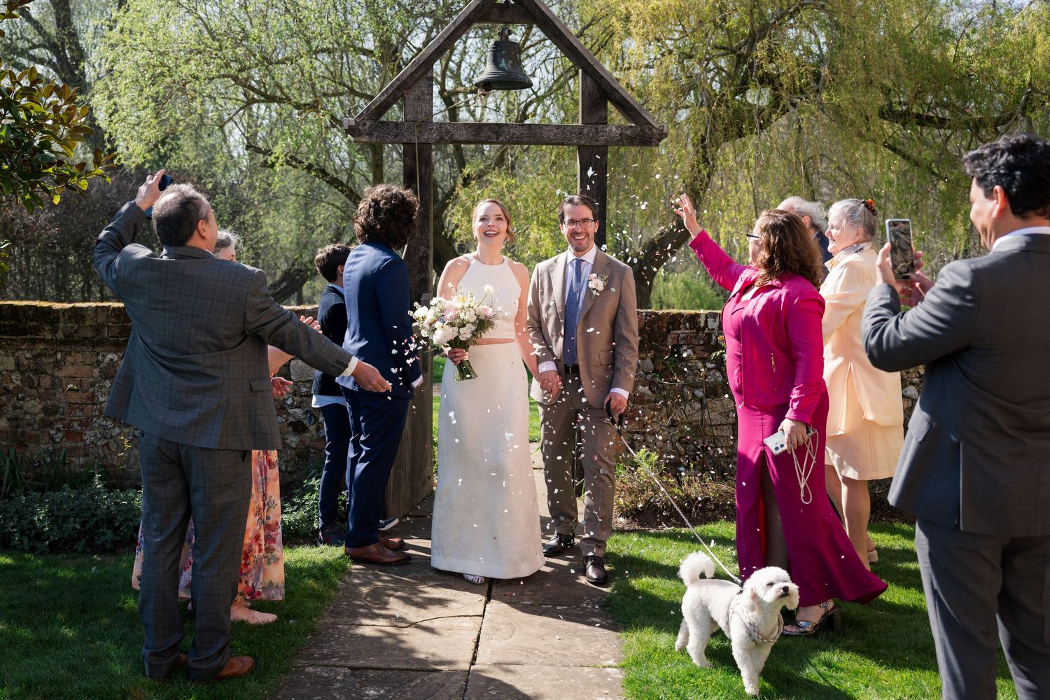 Bride and groom walk down outdoor aisle as guests celebrate with confetti at a sunny garden wedding ceremony.
