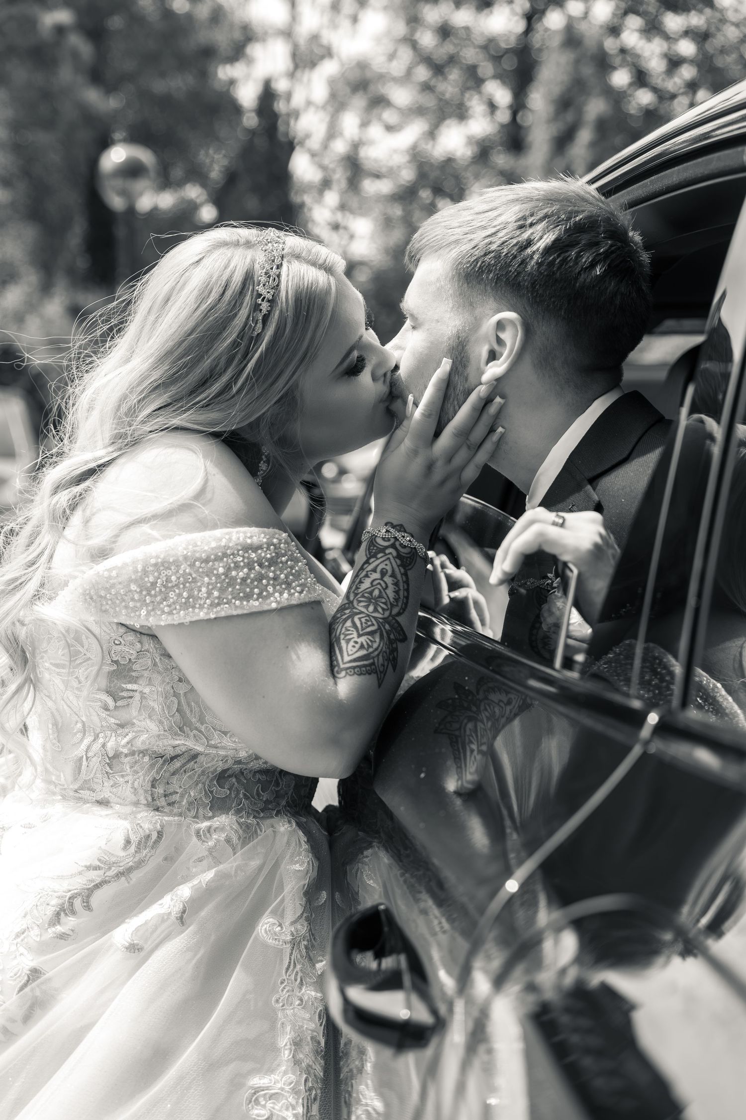 Black and white photo of a bride and groom sharing a romantic kiss beside a vintage car on their wedding day.