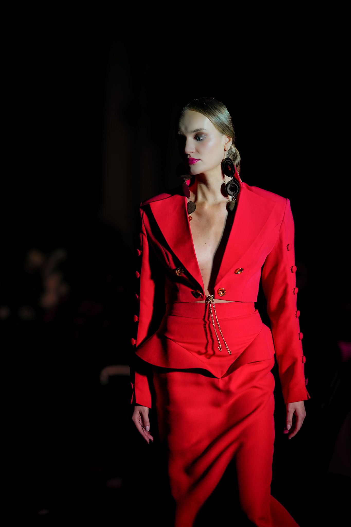 Model walks the runway in a striking red power suit with dramatic lighting against a dark background during fashion show.