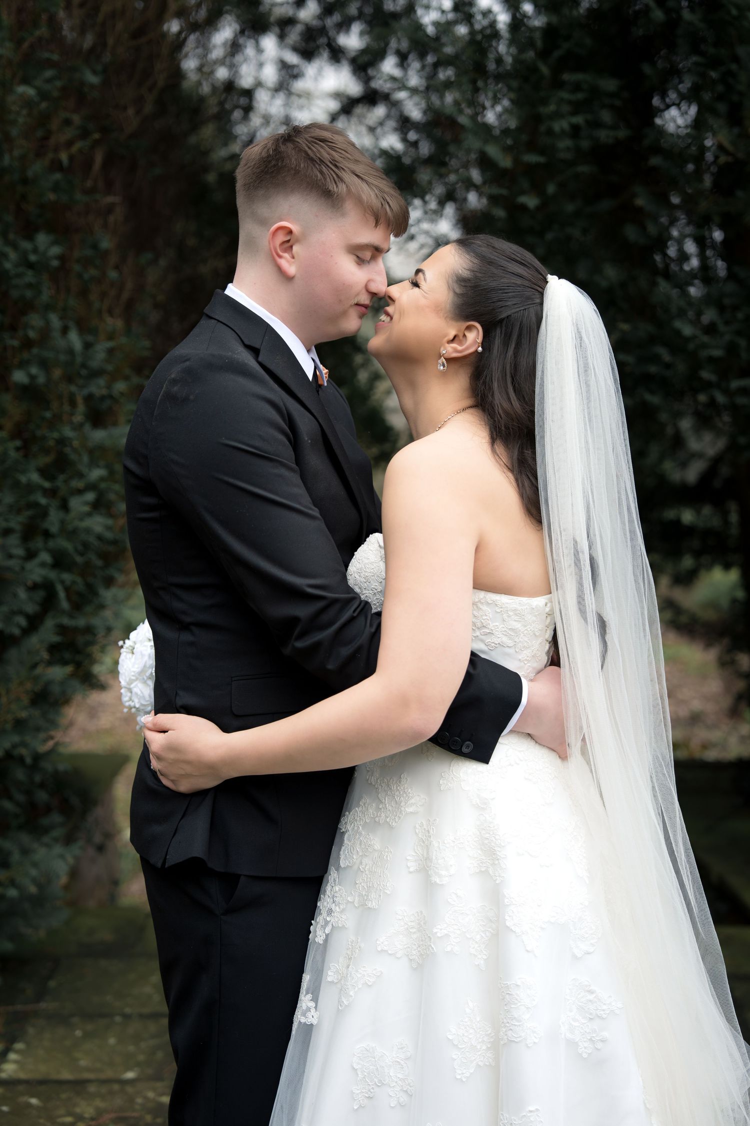 Bride and groom share a romantic almost-kiss moment outdoors, she in a white lace gown with veil, he in a black suit.