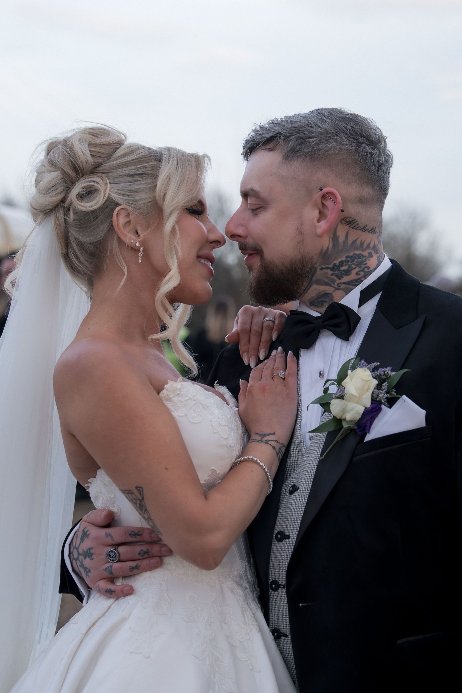 Bride and groom with tattoos share a romantic moment, dressed in wedding gown and black tuxedo with bow tie.