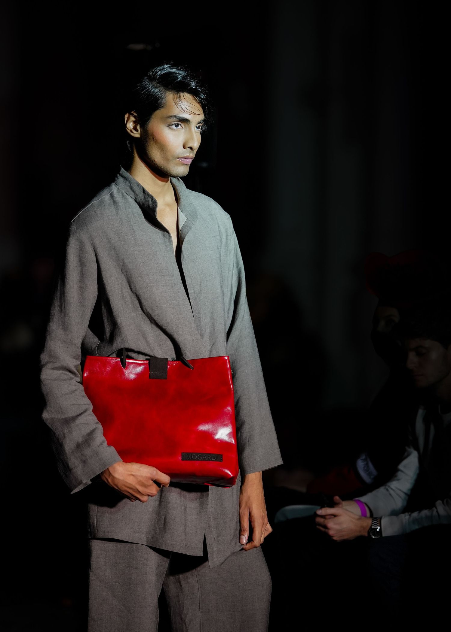 Model walks runway in gray suit with striking red patent leather belt during fashion show.