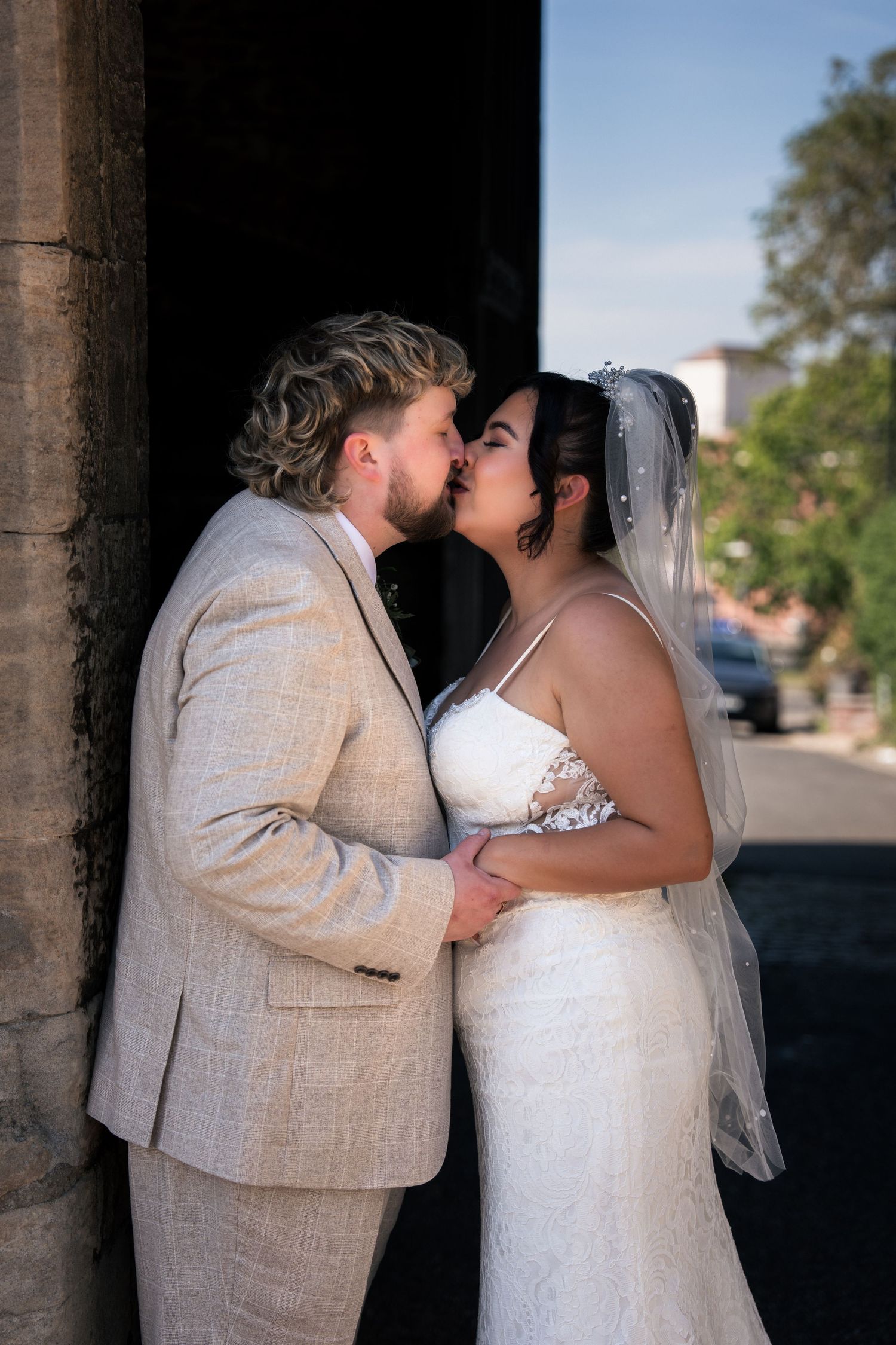 A bride and groom share a romantic kiss in wedding attire, with the bride in a white gown and veil outdoors.