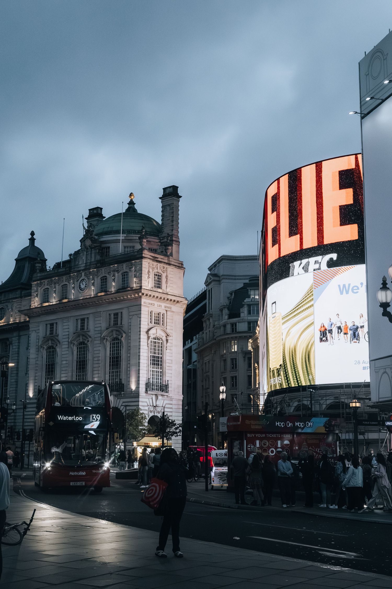 Stadsfoto van Piccadilly Circus bij avond, met verlichte reclameborden, stadsverkeer en drukke sfeer.