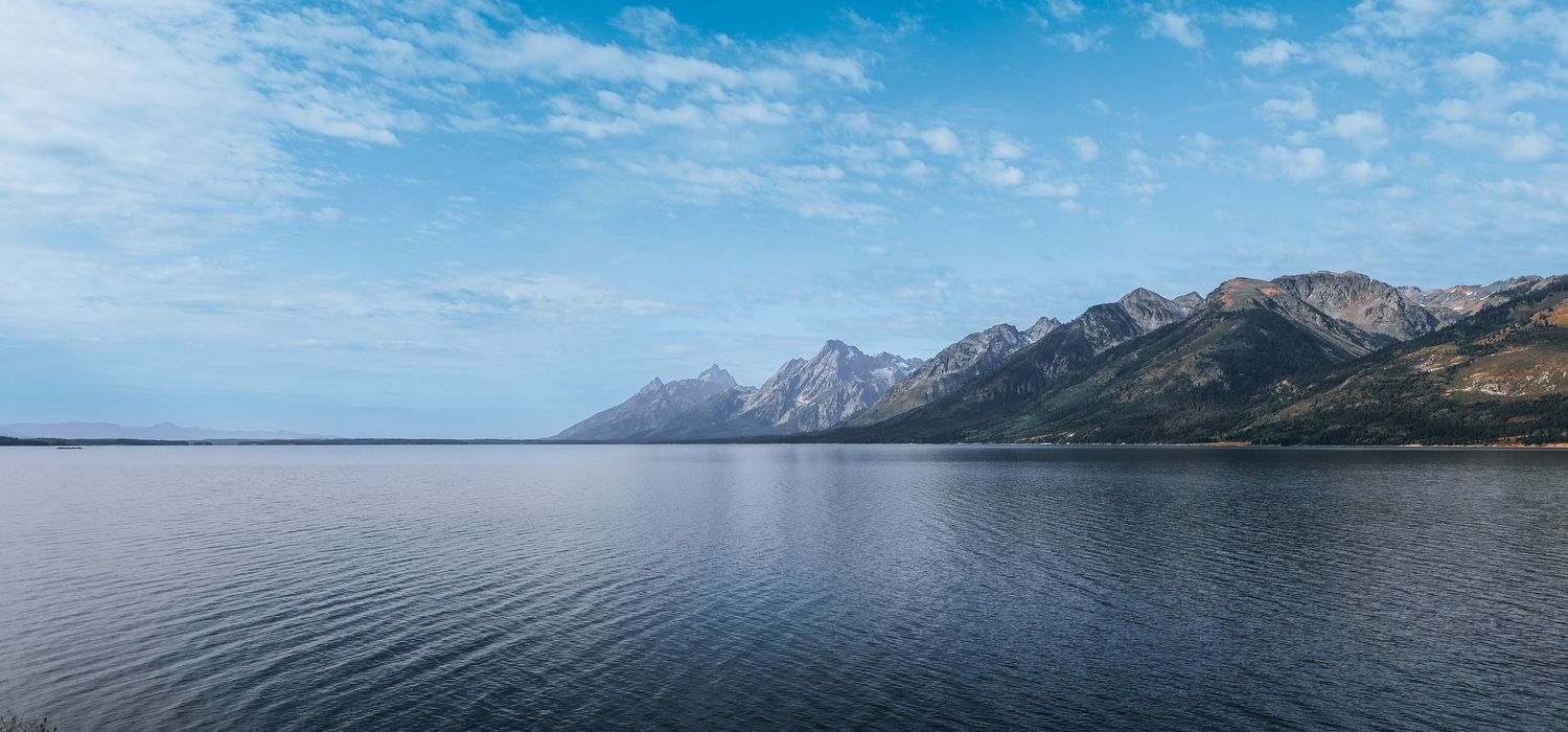 Landschapsfoto van een rustig meer met bergketen op de achtergrond, heldere lucht en kalme reflecties in het water.