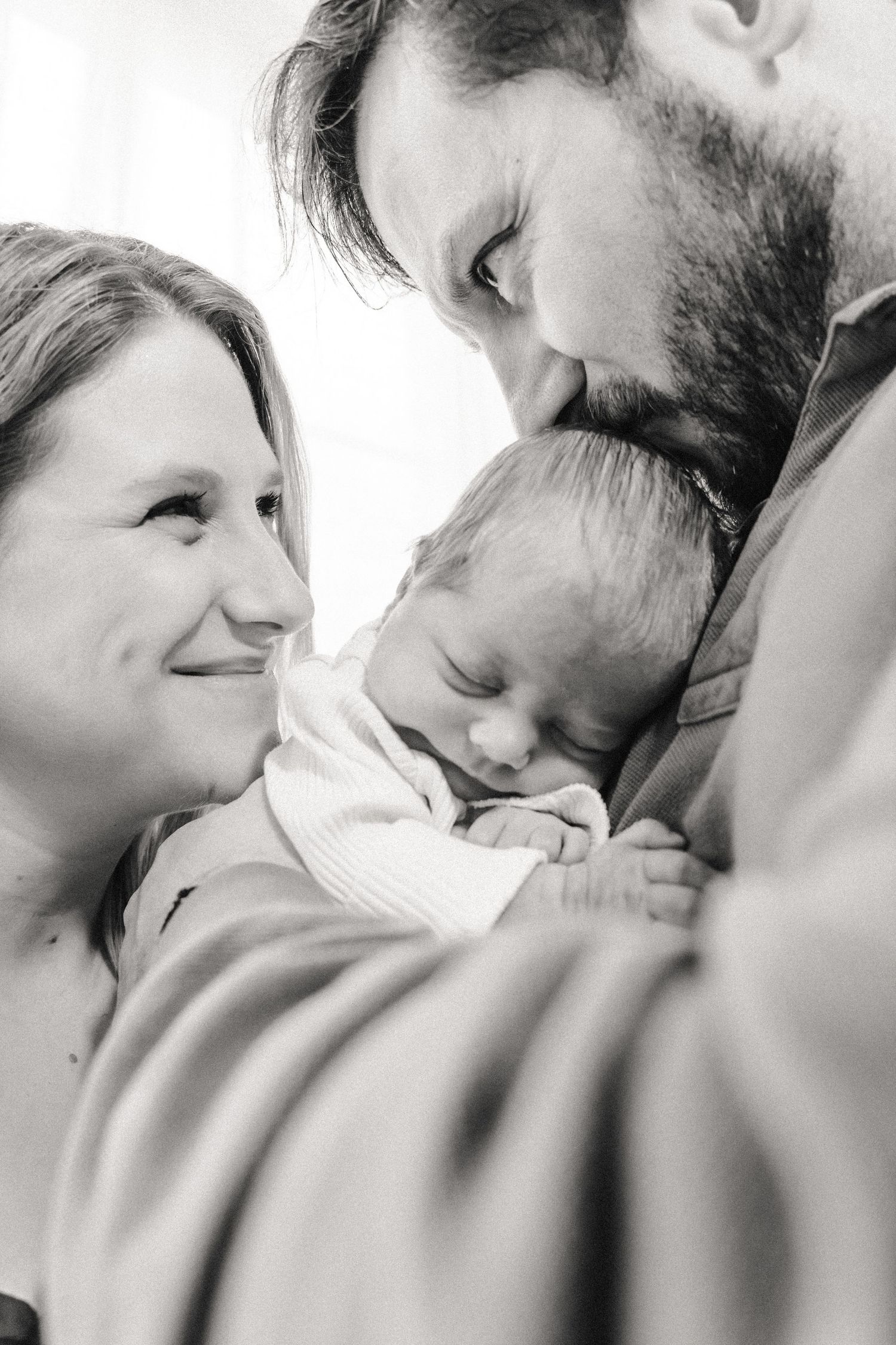 New parents cuddle newborn baby in well-lit room during at-home newborn session