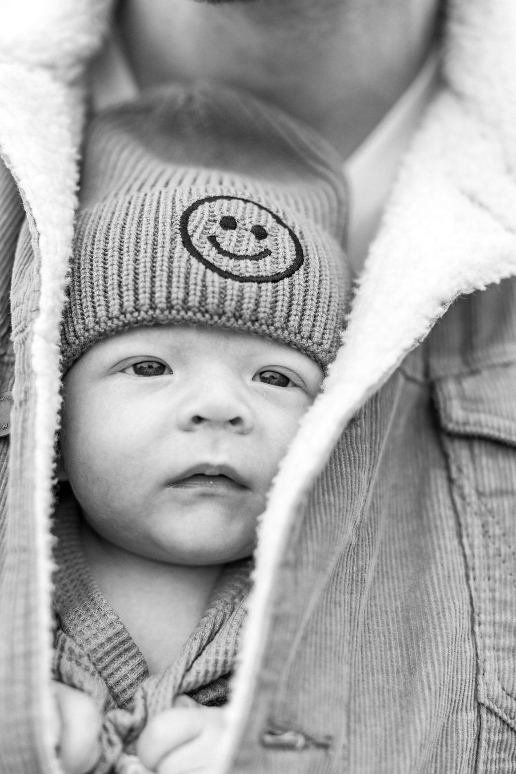 Newborn baby with smiley-face hat snuggled in dad's jacket
