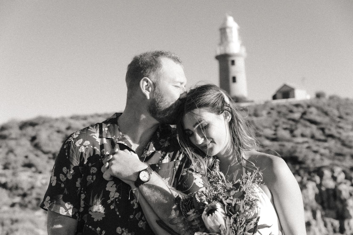 Man kisses forehead of woman with lighthouse in the background | beach elopement photography