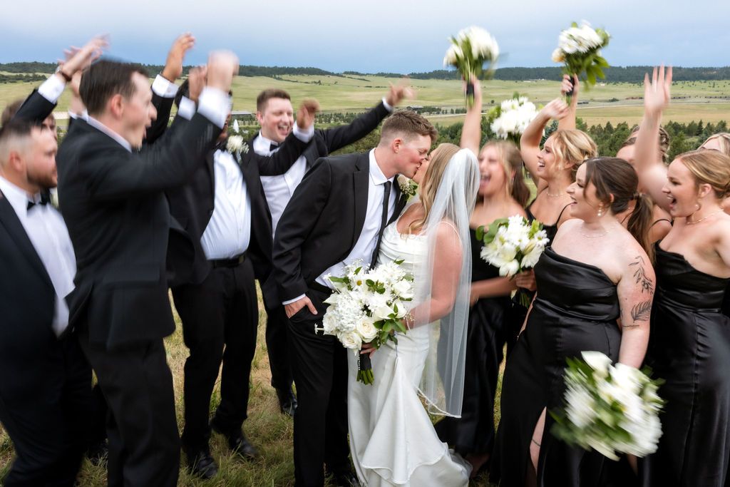 Newlyweds kiss surrounding by celebrating wedding party in Colorado field | Colorado Wedding Photography | Spruce Mountain