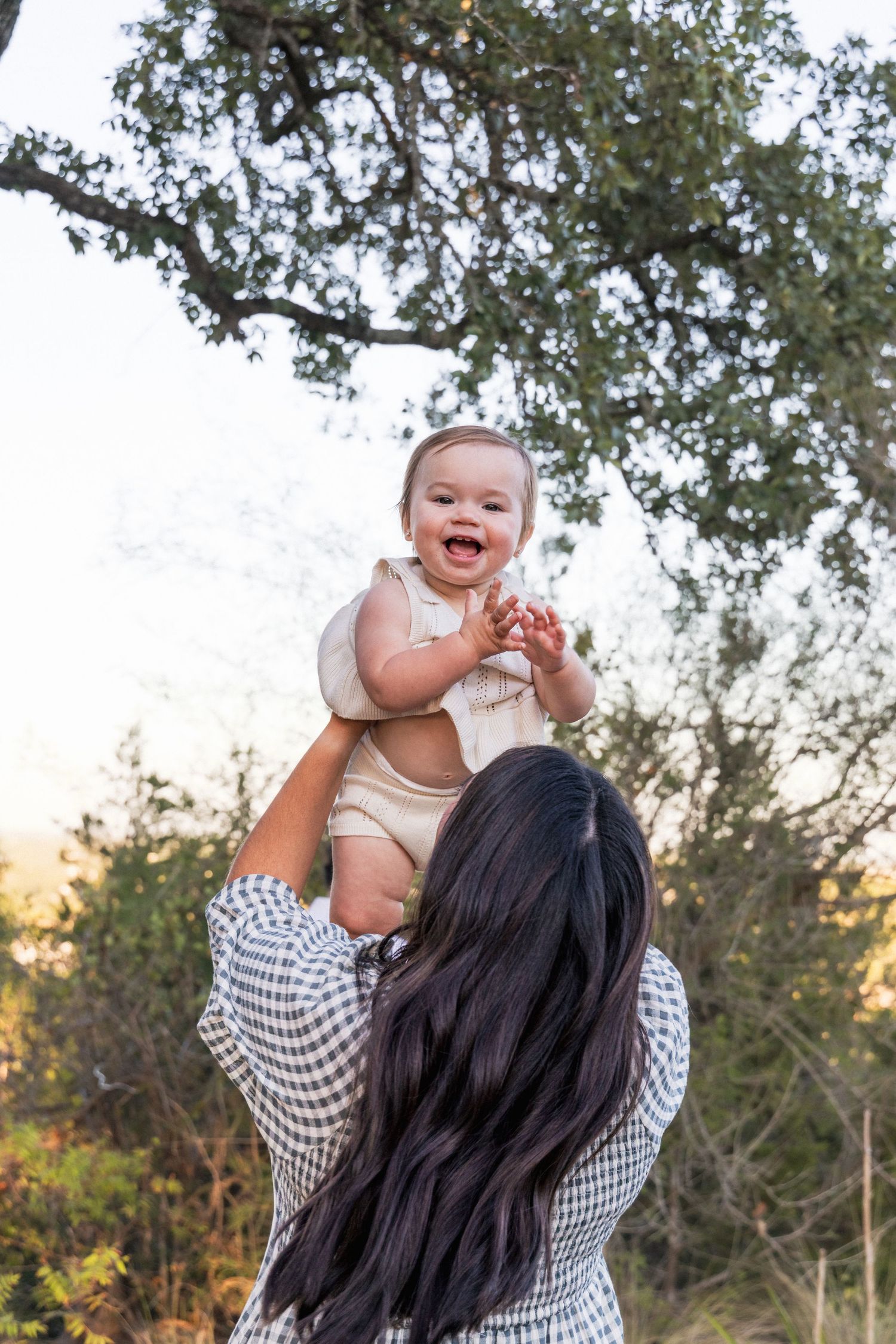 Mom holds infant over her head and baby laughs surrounded by oak trees