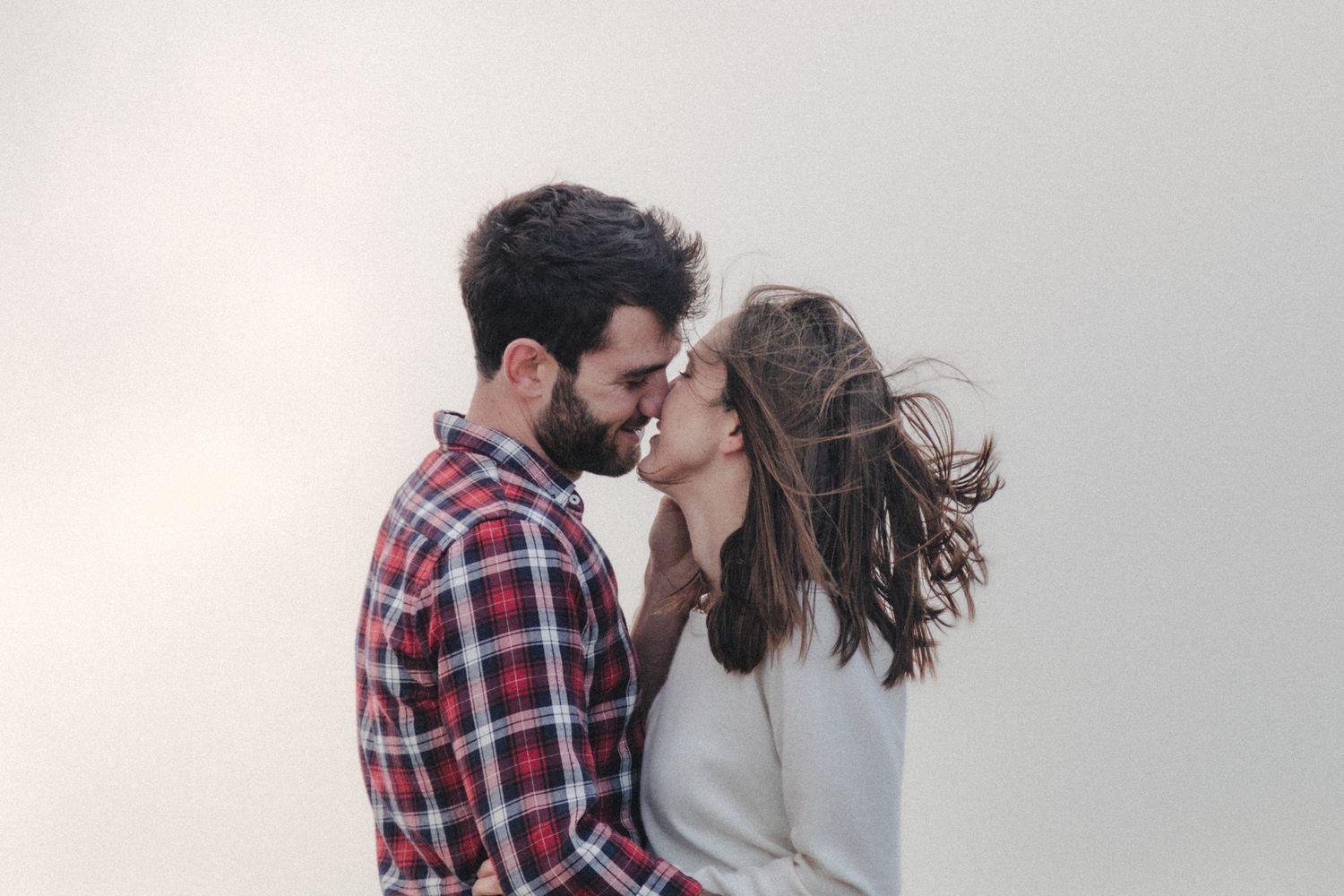 Couple embraces and kisses with wind-blown hair and fog during coastal engagement session