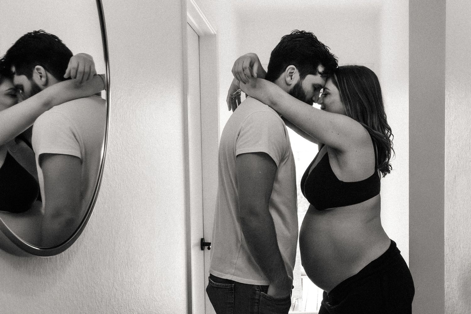 Pregnant woman embraces partner in hallway during at-home maternity photography session in black and white