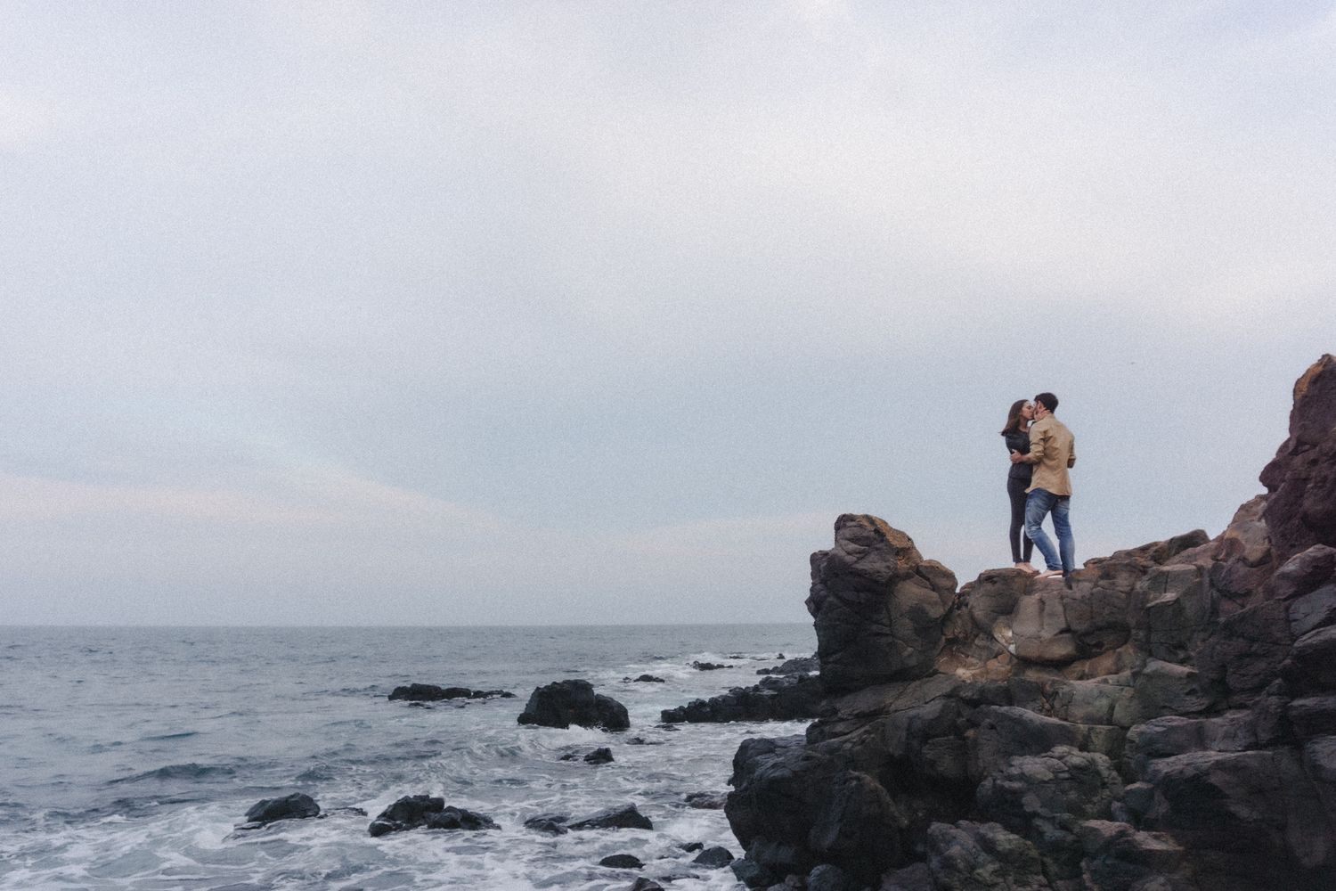 Barefoot couple embraces on black rocks during moody coastal engagement session
