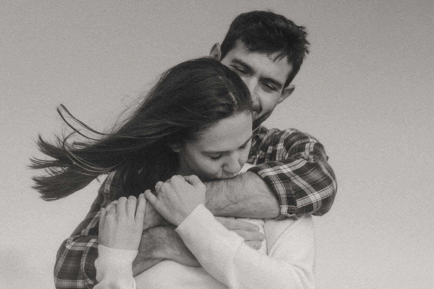 Black and white edit of woman kissing partner's arm with wind-blown hair during foggy coastal engagement session