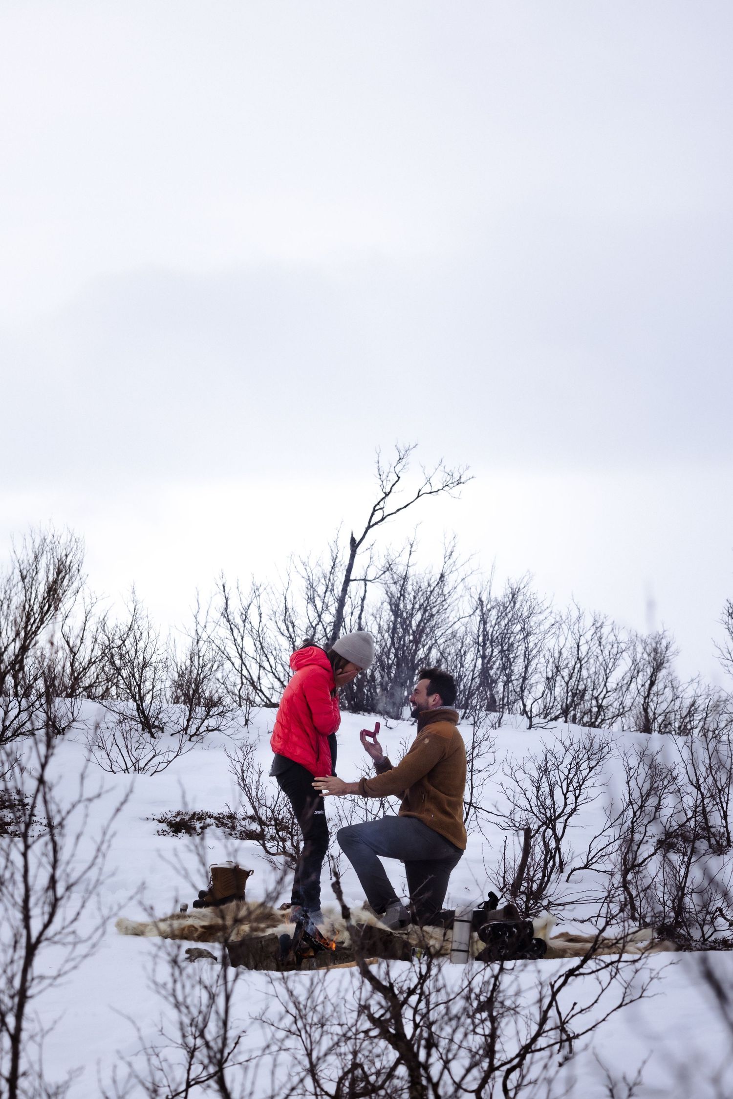 Couple in winter gear getting engaged by an outdoor fire in Arctic Sweden.