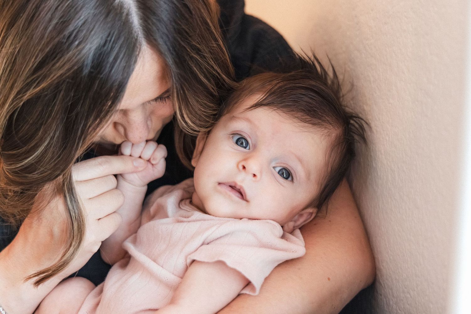Mother holding her baby near a sunlit window and kissing the baby's hand