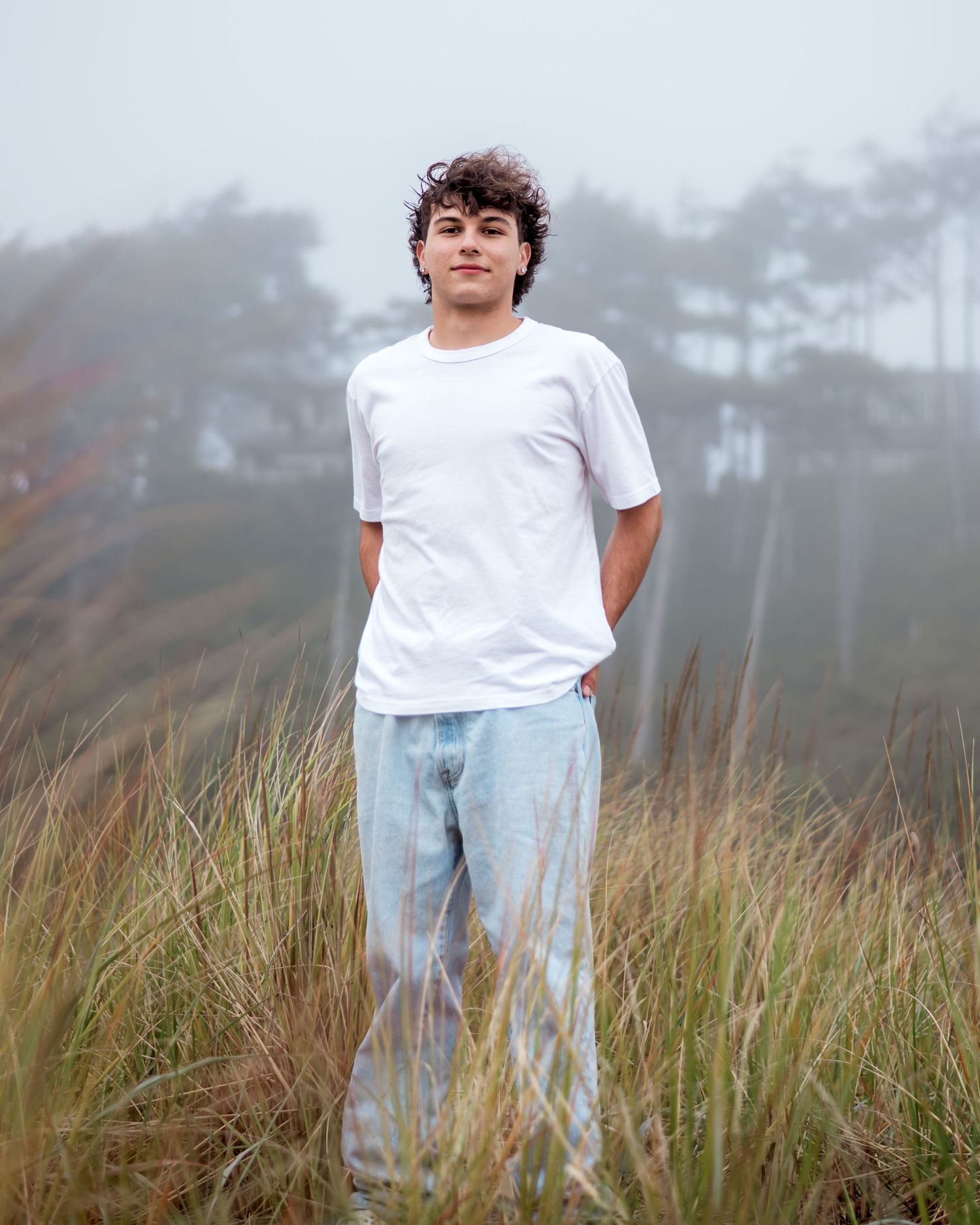 Person in white clothing stands in tall grass field on misty morning with foggy trees in background.