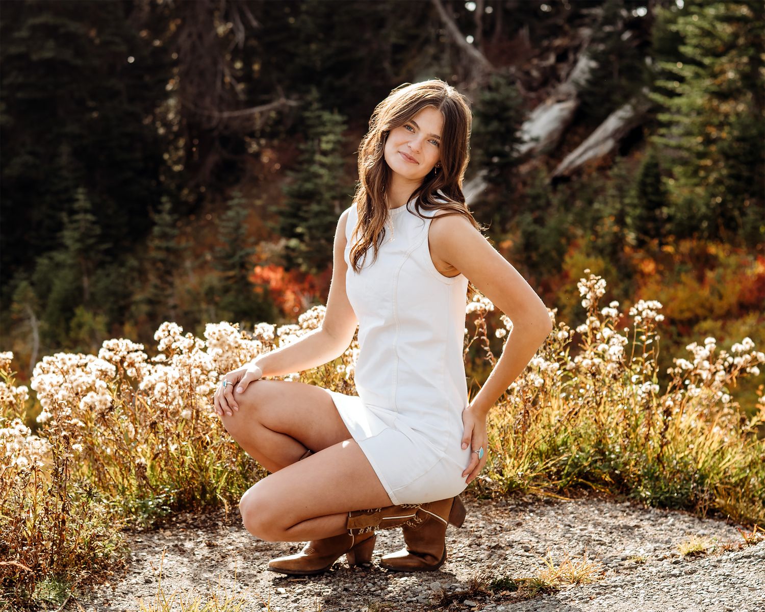 Young woman in white dress poses gracefully among wildflowers with autumn foliage and pine trees in background.