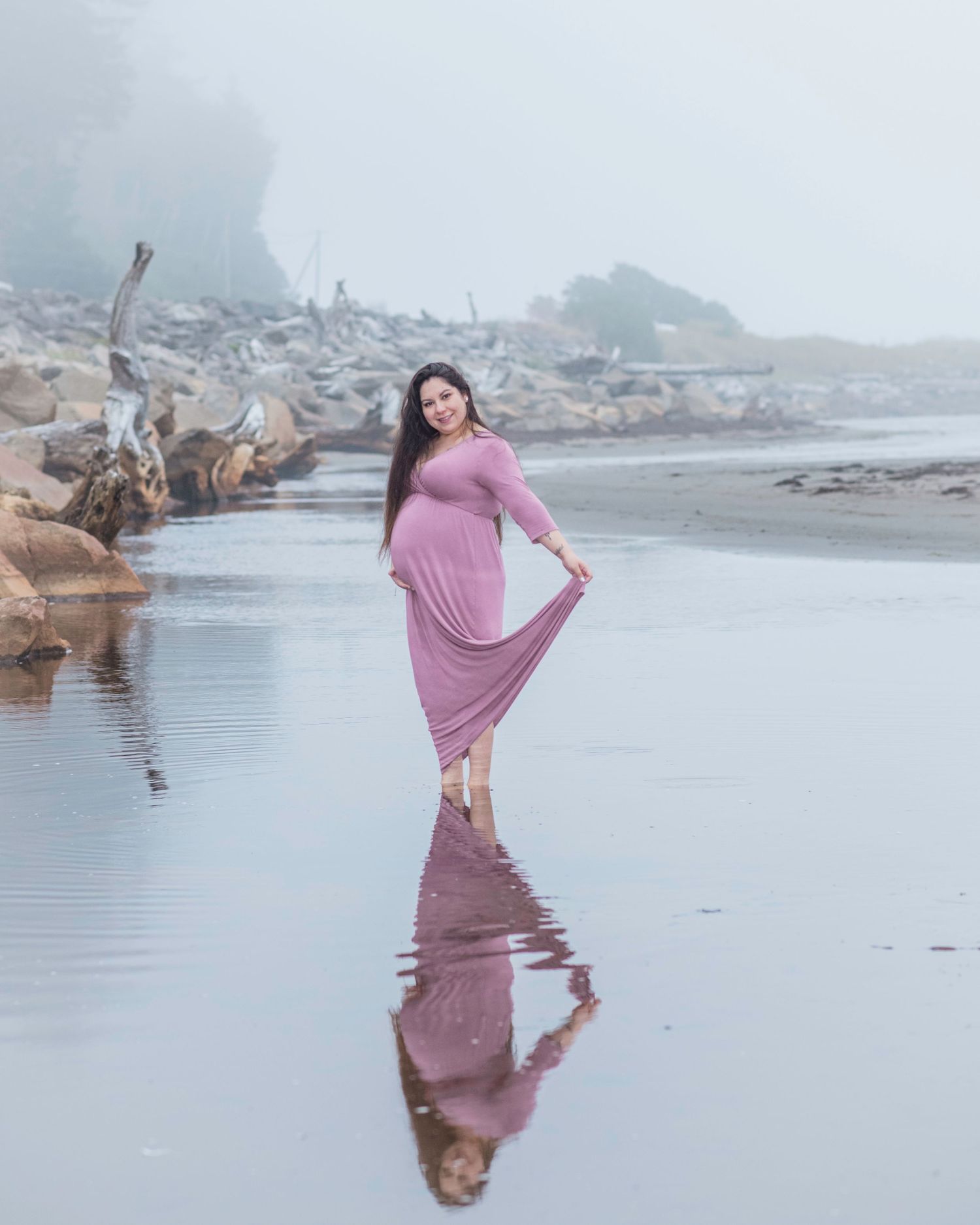Woman in flowing pink dress poses gracefully on misty beach with driftwood.