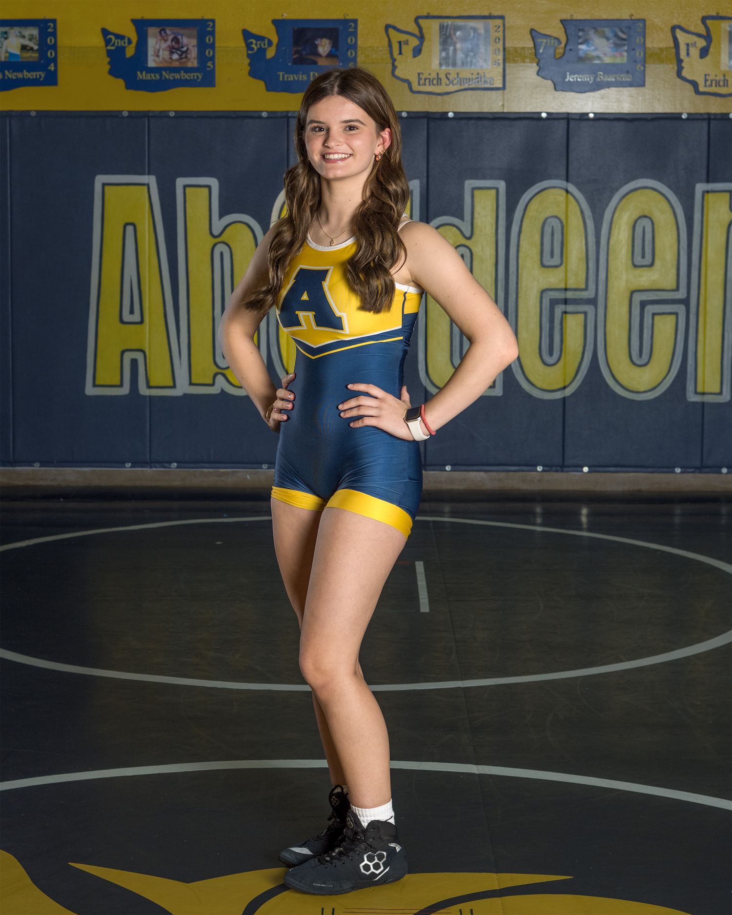 Student athlete poses in blue and gold wrestling uniform on school mat.