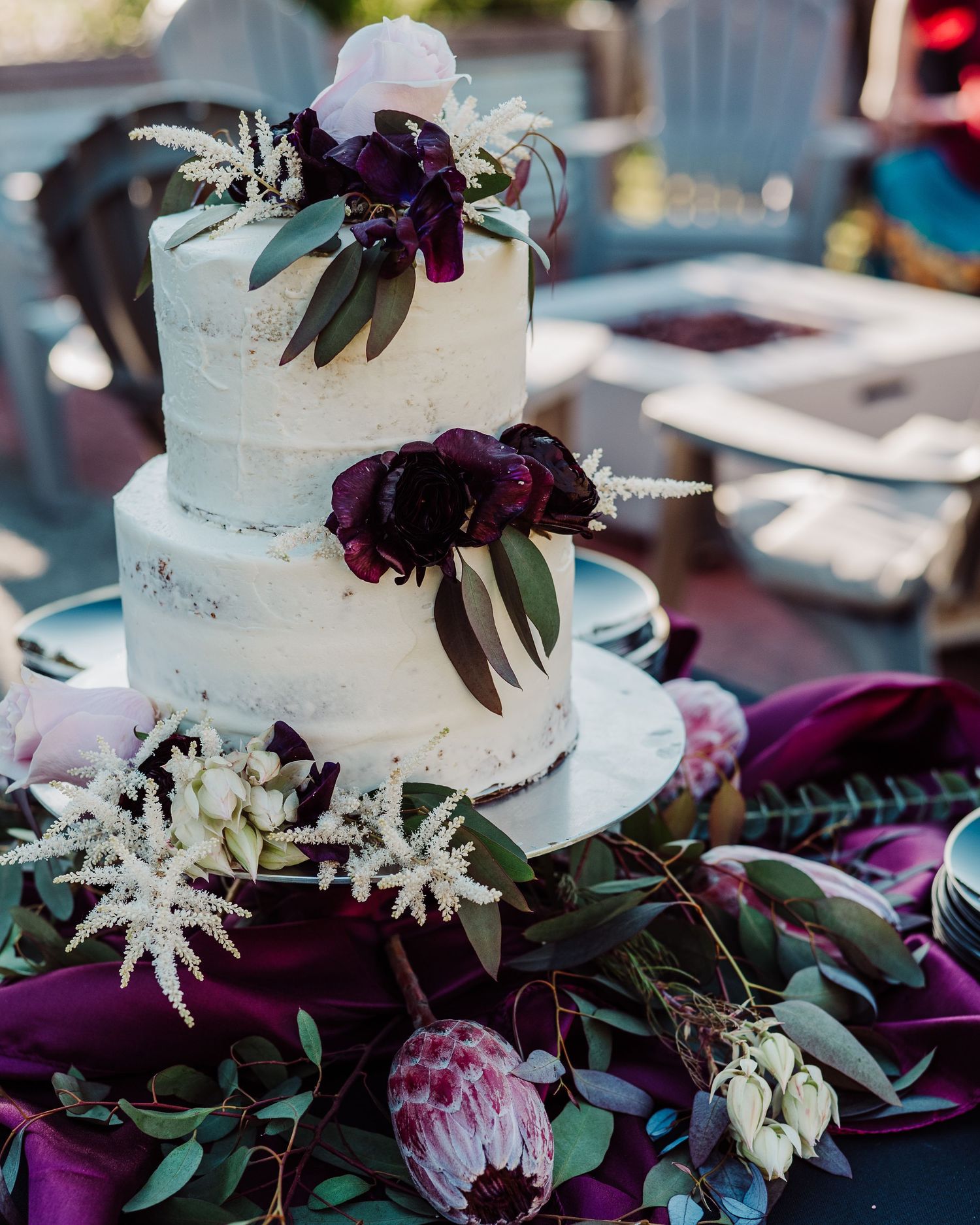 Elegant white wedding cake decorated with deep purple flowers and greenery on a dark tablecloth.